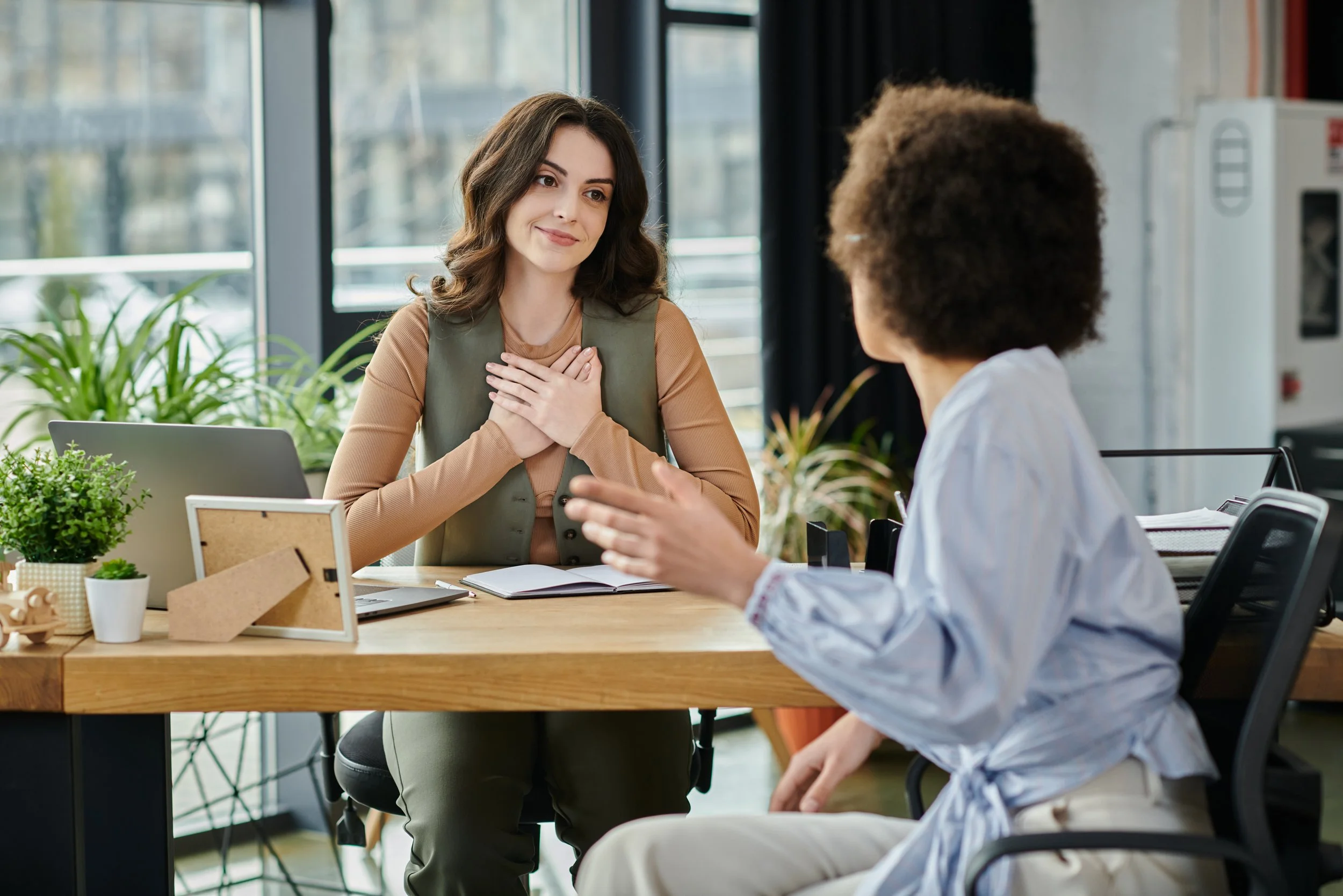 Mujer sonriendo en una entrevista de trabajo, con las manos en el pecho, en una oficina moderna con plantas y ventanas grandes.