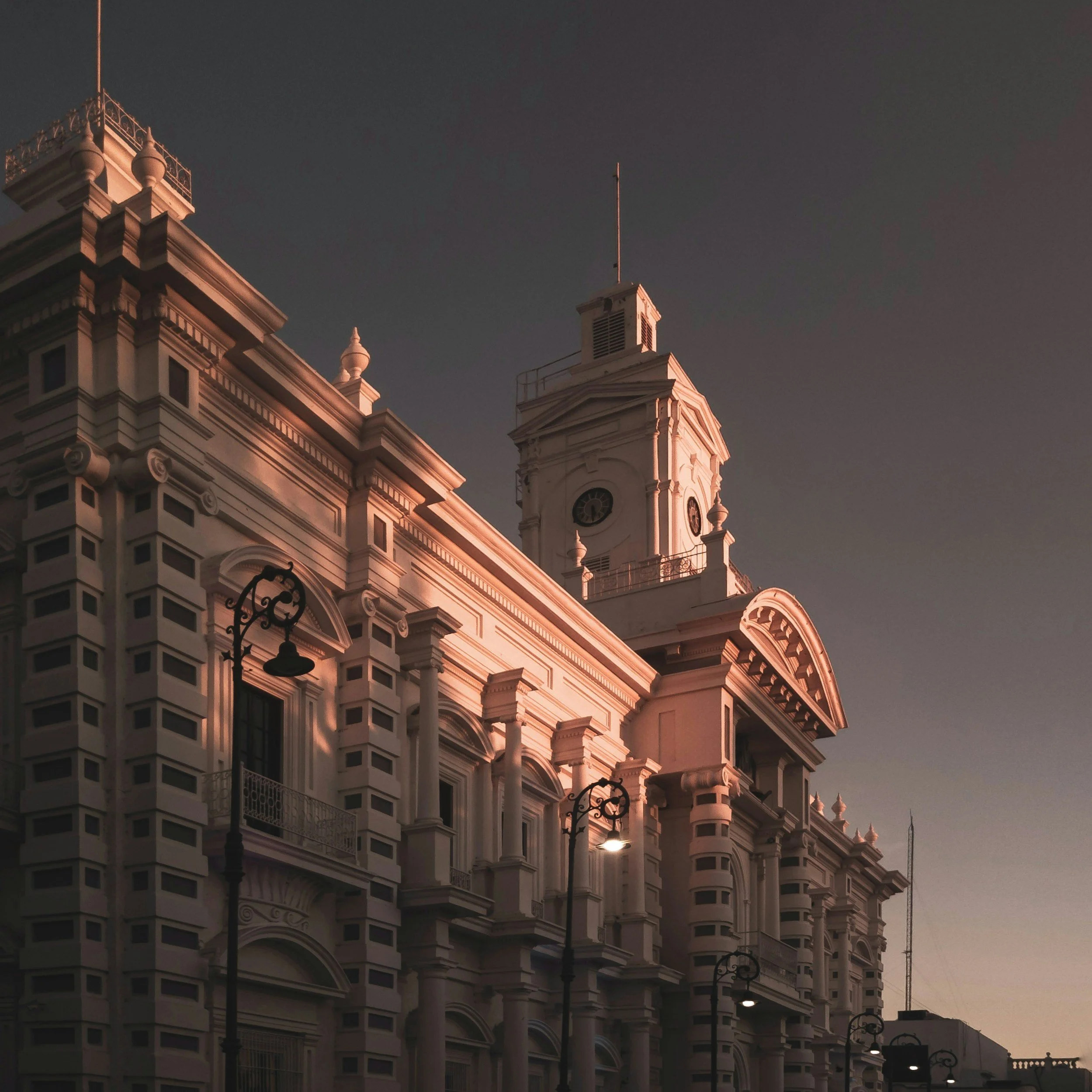 Edificio histórico con detalles arquitectónicos ornamentales, reloj en la torre y farolas en primer plano, al anochecer.