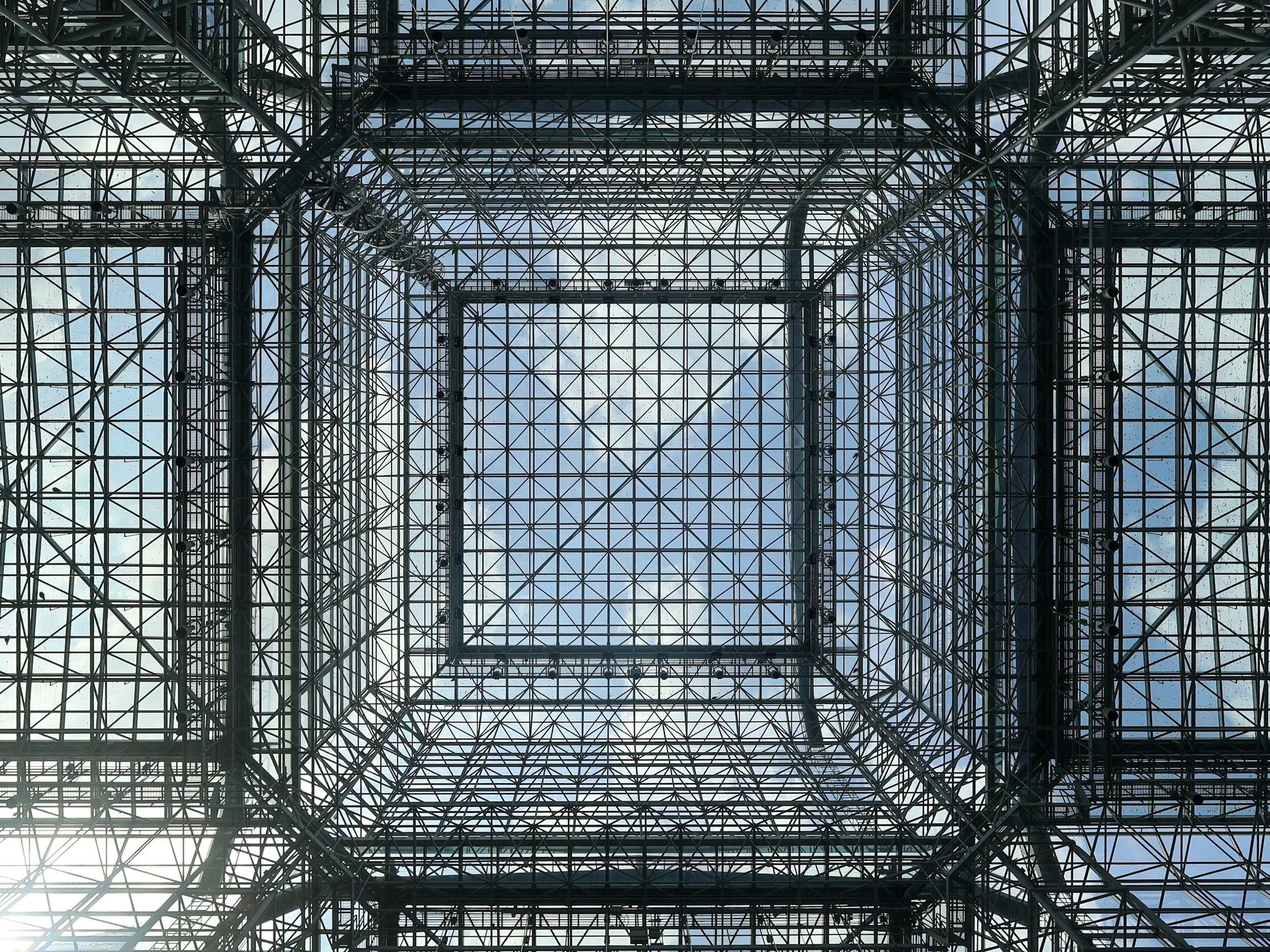 Vista hacia arriba de una estructura de acero y vidrio formando un cubo con el cielo en el fondo.