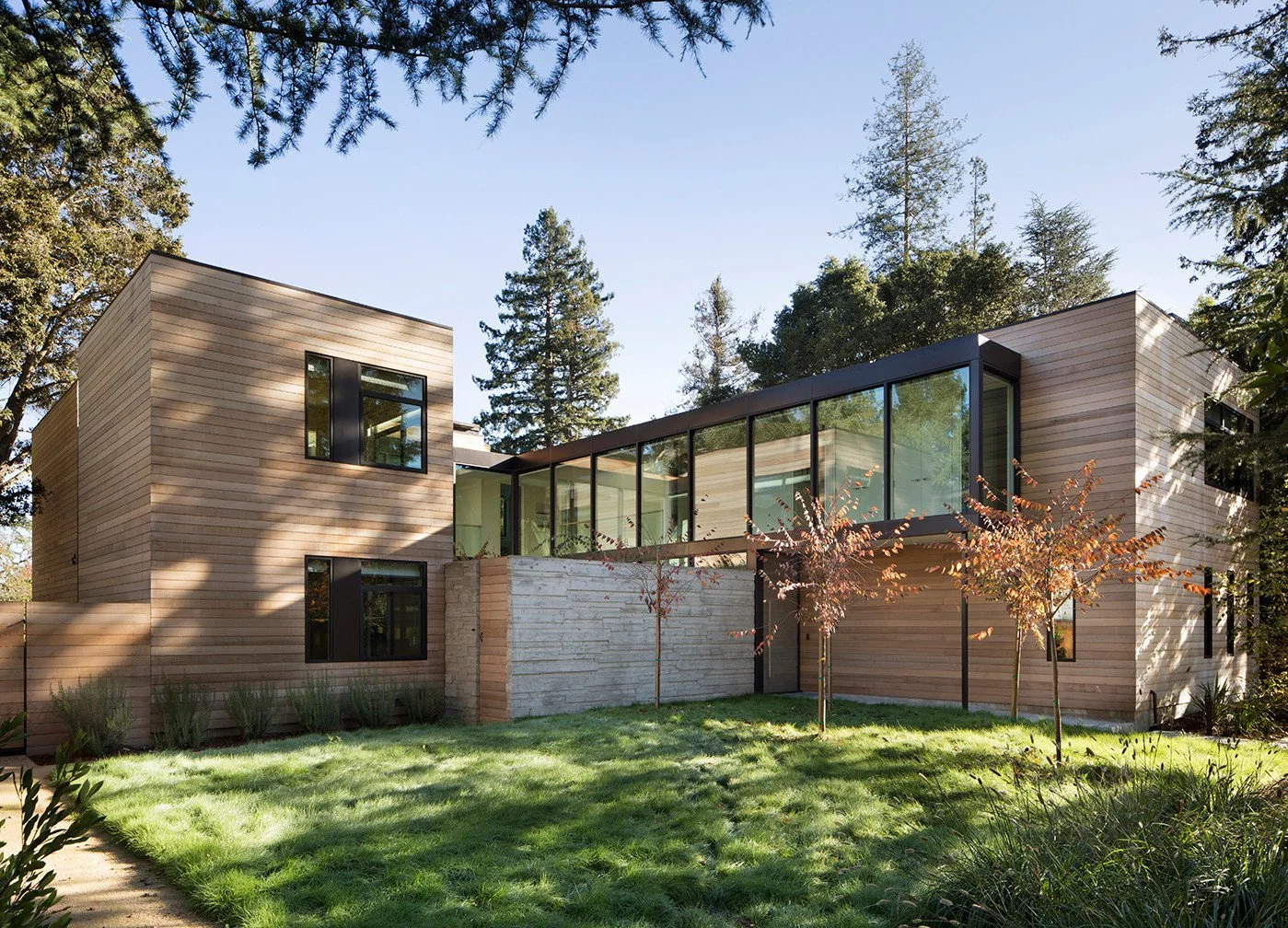 Modern house with large glass windows and wood siding, surrounded by a green lawn and tall trees, under a clear blue sky.