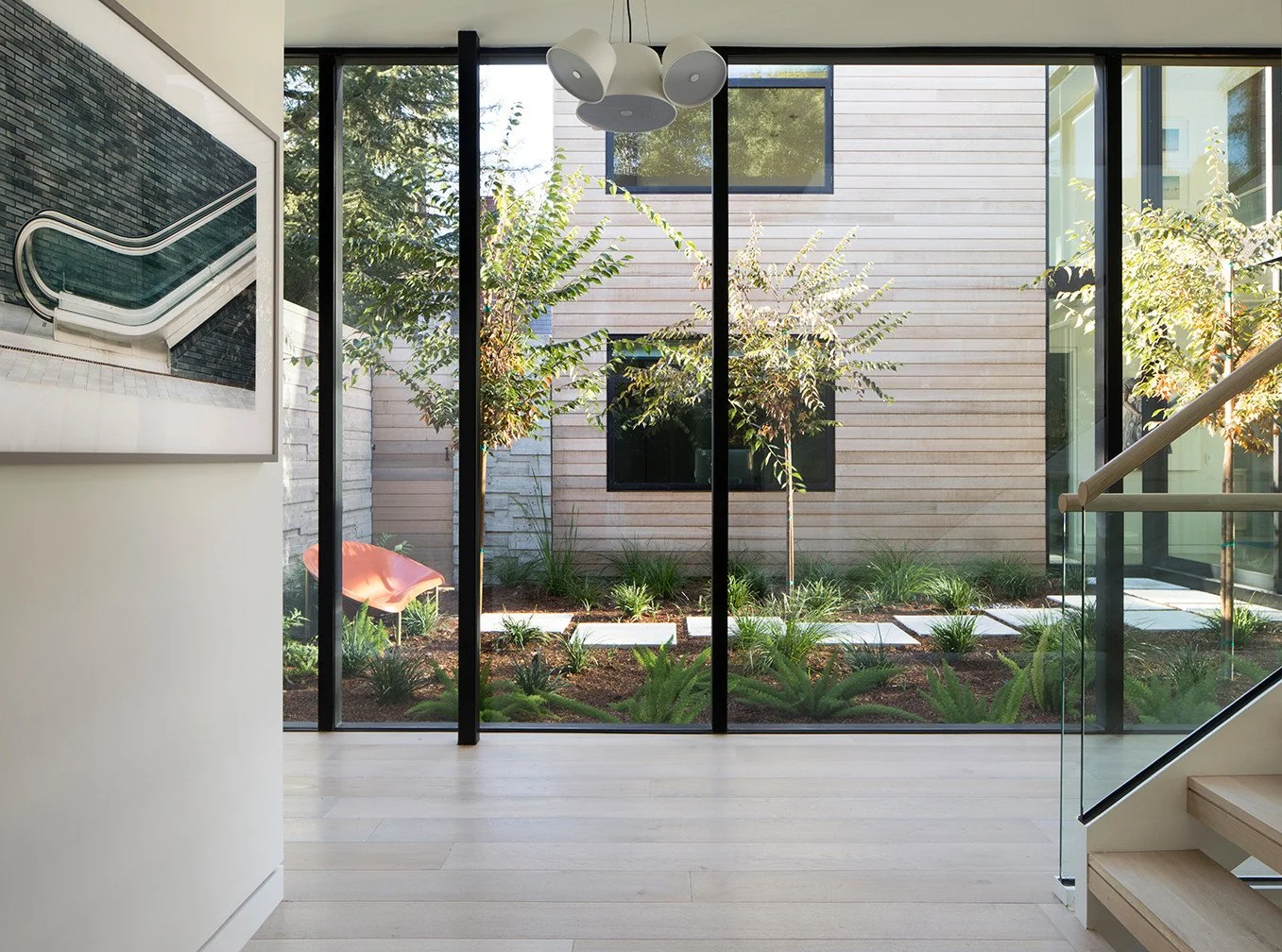 Modern interior space with wooden flooring, large floor-to-ceiling glass windows, view of a small garden with trees, plants, and a pink chair outside, and a staircase with glass railing on the right side.