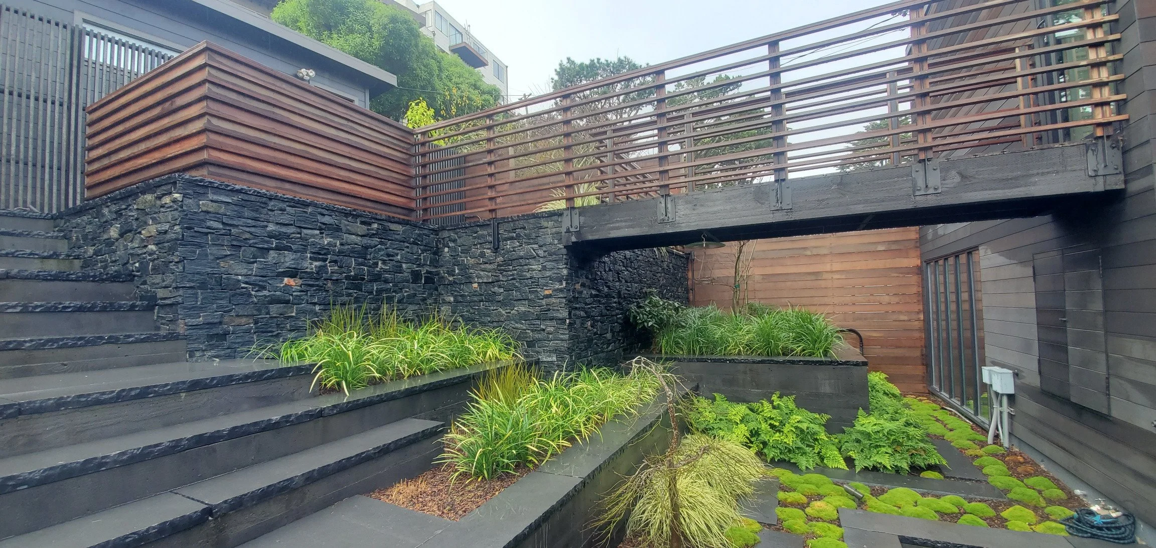 Modern backyard with black stone and wood accents, featuring raised garden beds with green plants and a small water feature, with stairs and a wooden deck.