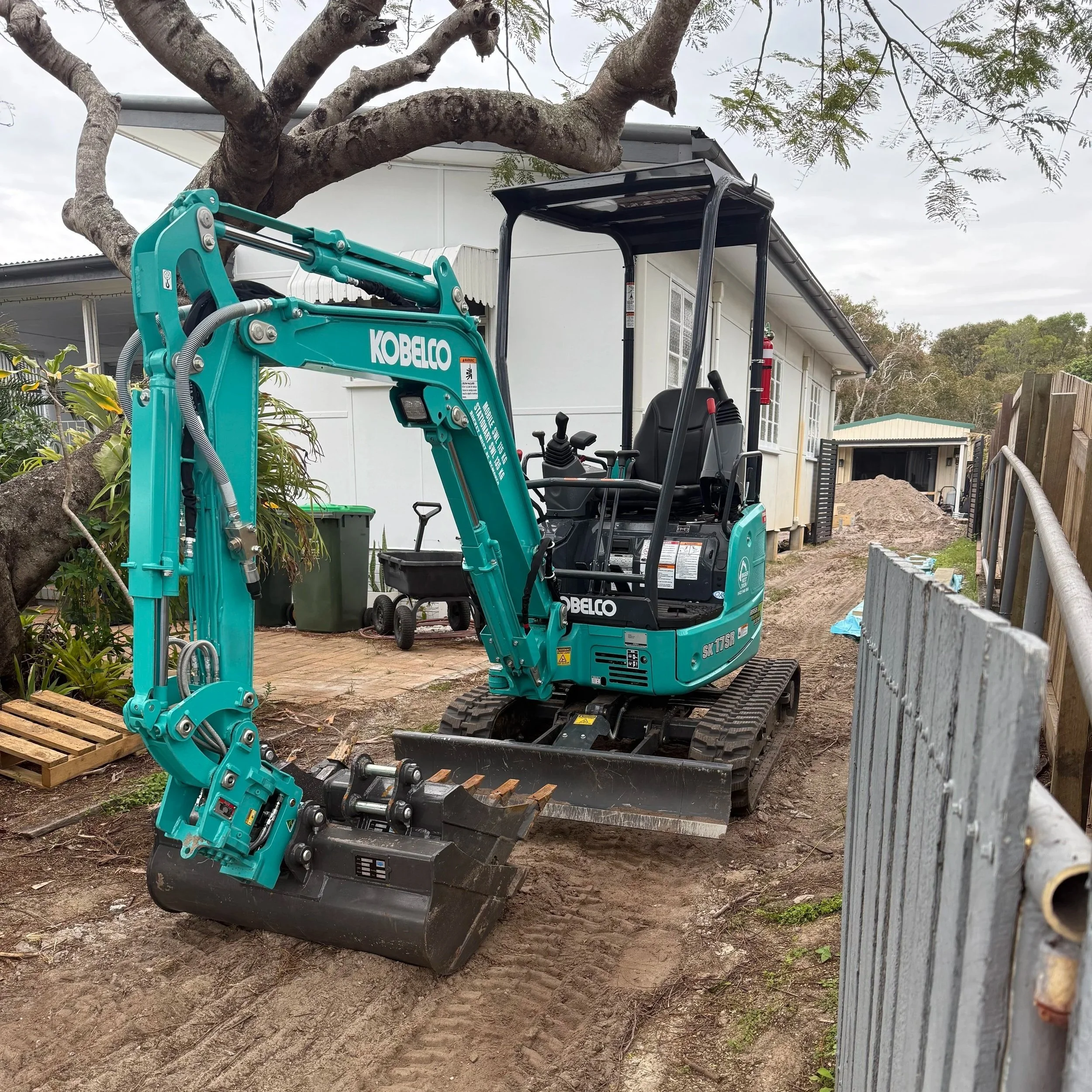 A teal-colored Kobelco mini excavator parked on a dirt path at a construction site, with a tree and house in the background.
