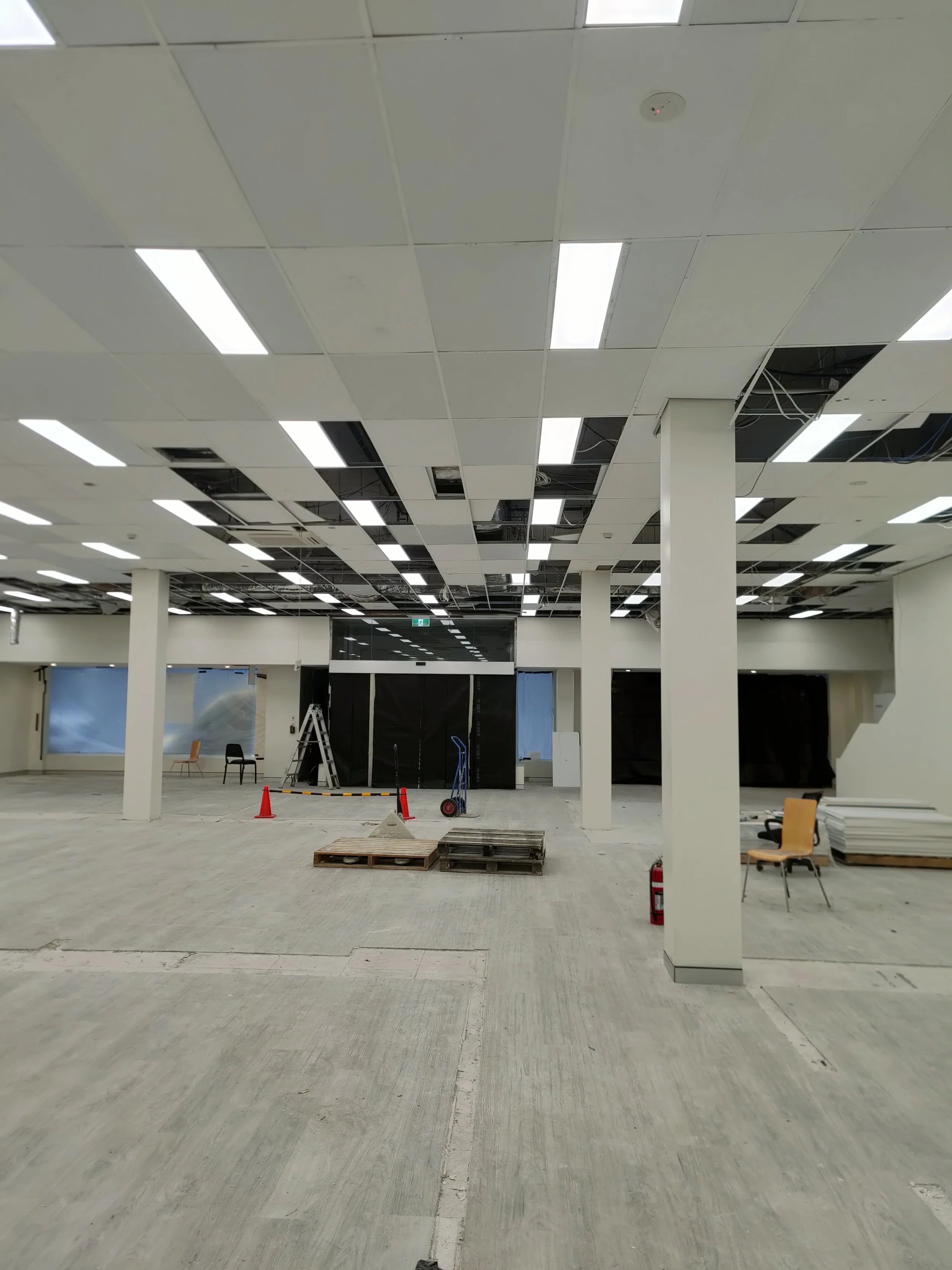 Interior of a commercial space under renovation with exposed ceiling tiles, construction equipment, and some chairs and pallets.