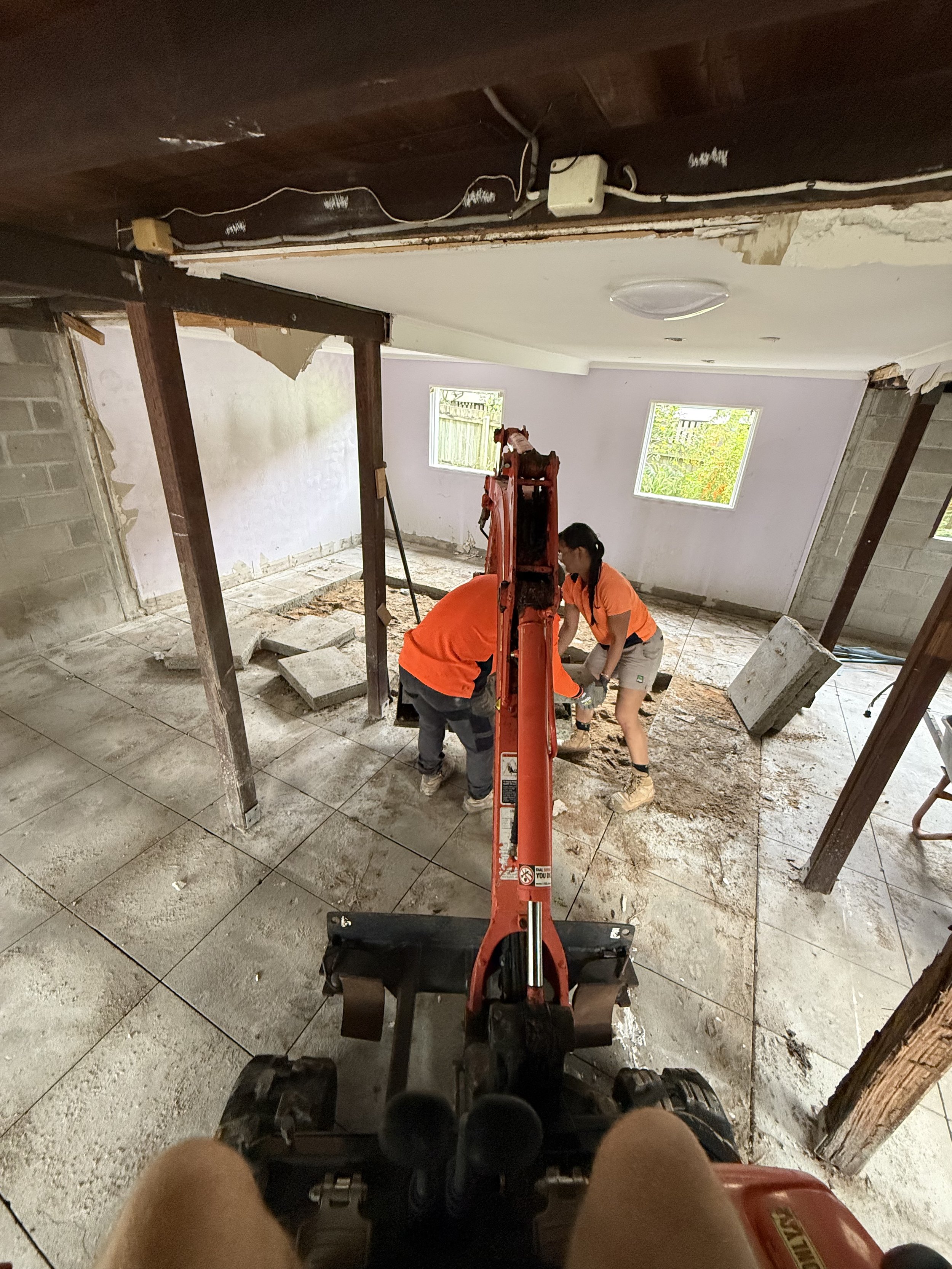 Two construction workers are working in a room with exposed drywall and concrete floor. They are using a backhoe to dig and move dirt or debris. The room has two small windows, and one section of the wall is exposed cinder blocks. There are wooden supports and construction materials around.