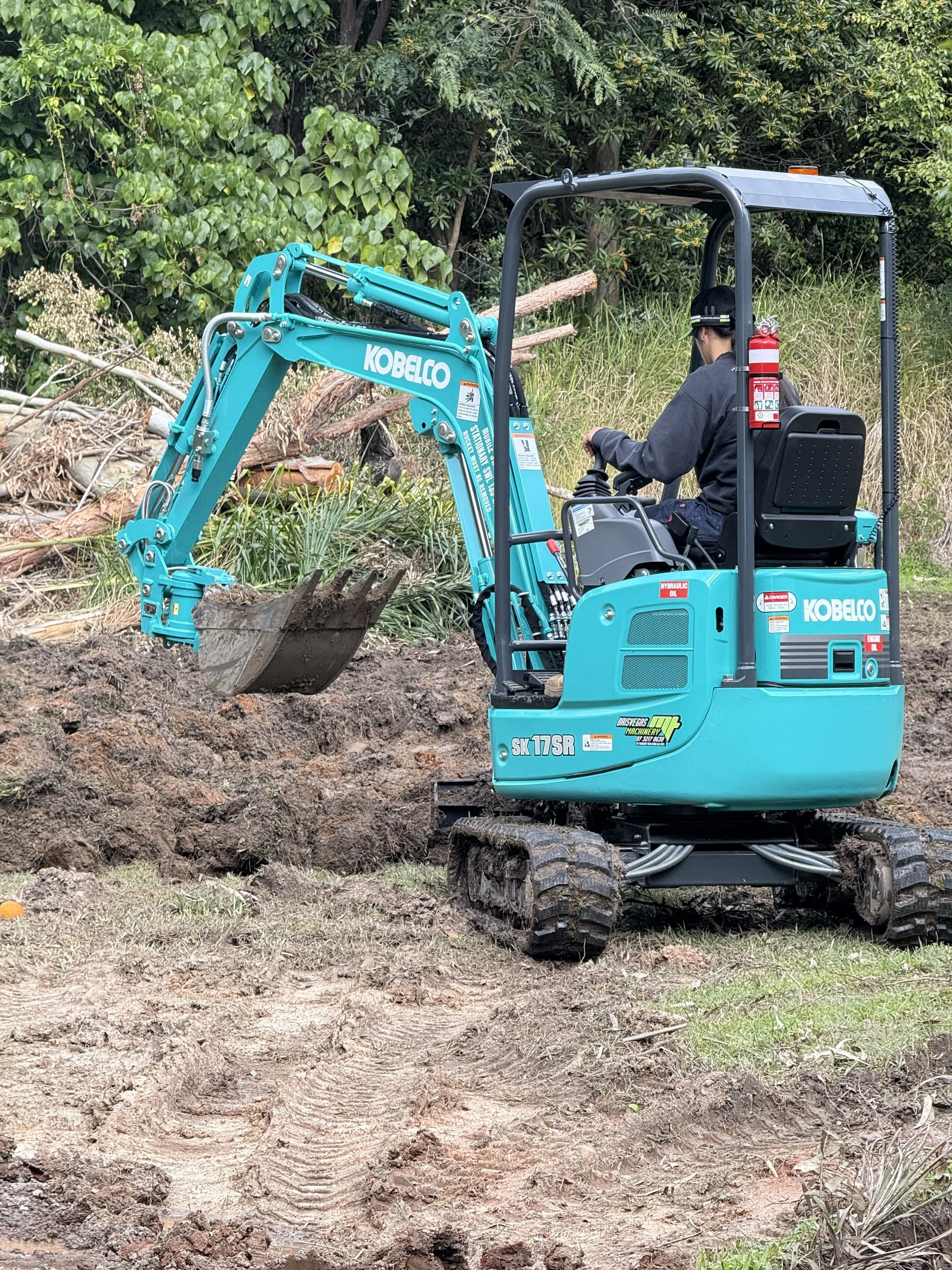 A person operating a blue Kobelco mini excavator on a dirt construction site surrounded by trees and greenery.