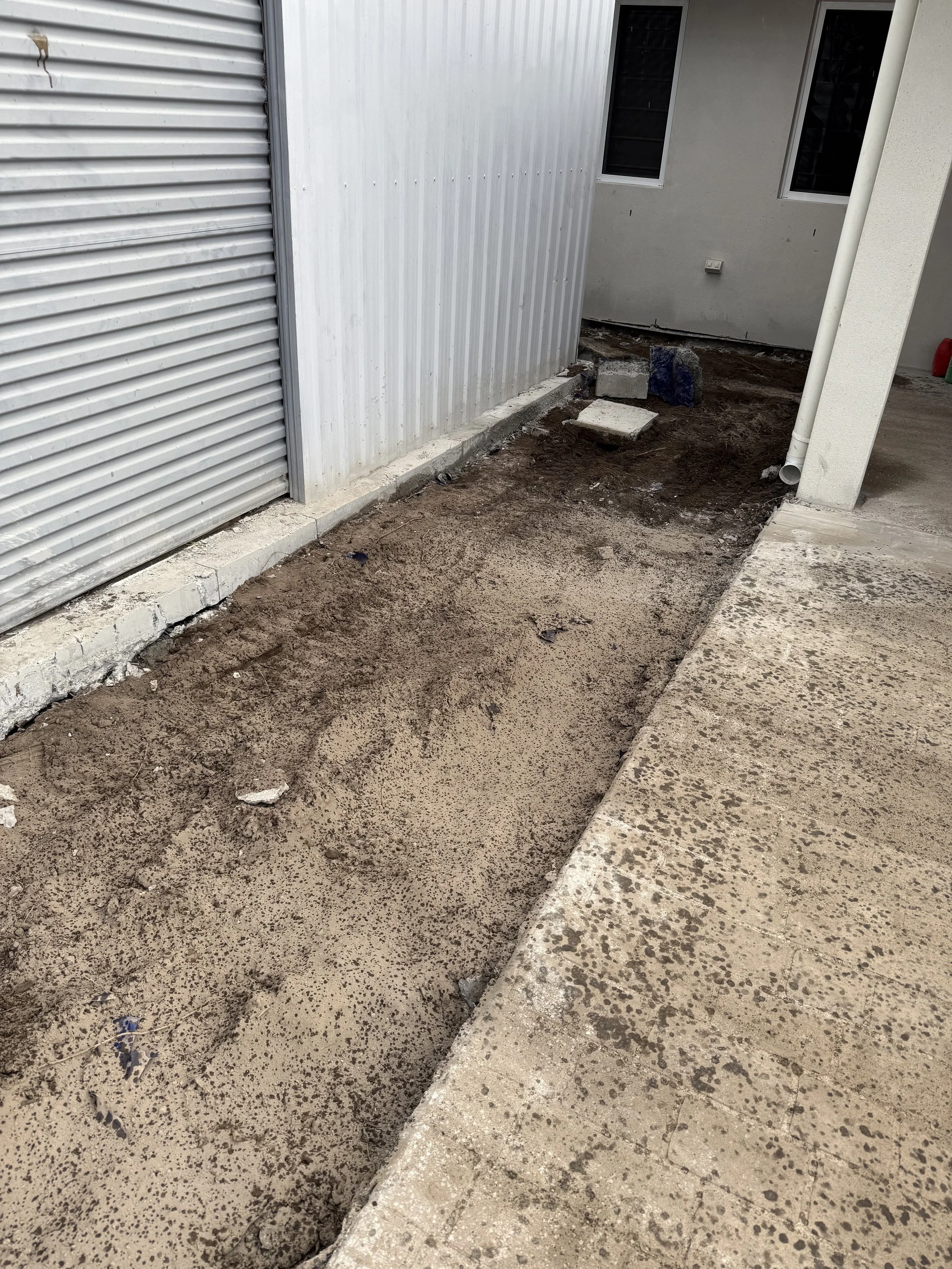 Concrete removal on a construction site with dirt and concrete on the ground, partially paved sidewalk, and building walls with windows and metal siding.