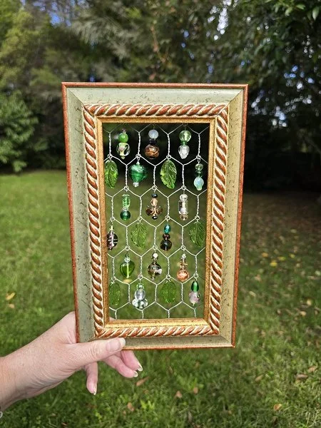 Hand holding a decorative mirror frame with hanging beads and shells, outdoors in a grassy area with trees in the background.