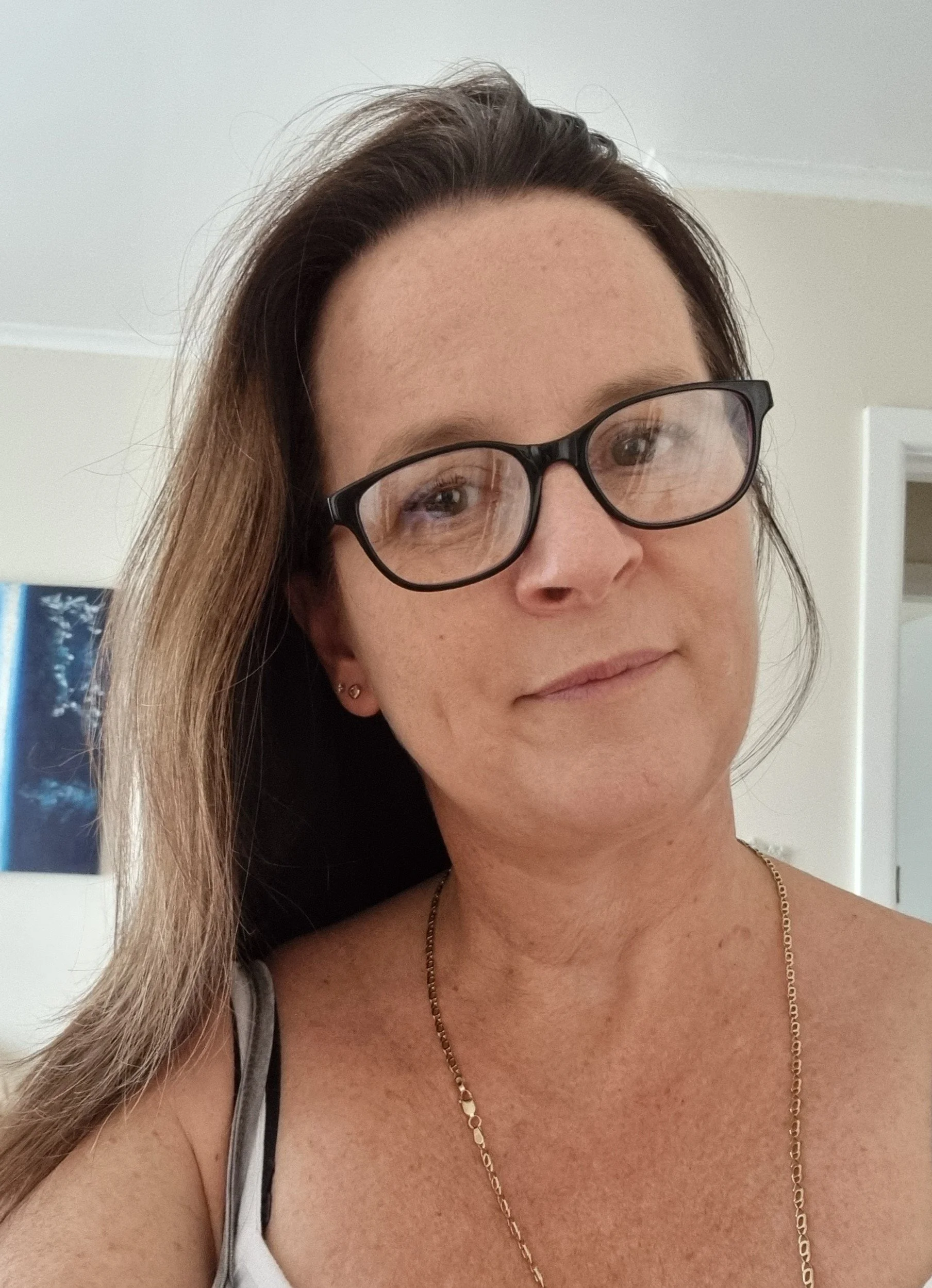 Close-up of a smiling woman with long brown hair, black glasses, and gold jewelry, standing indoors with a white wall and a door in the background.