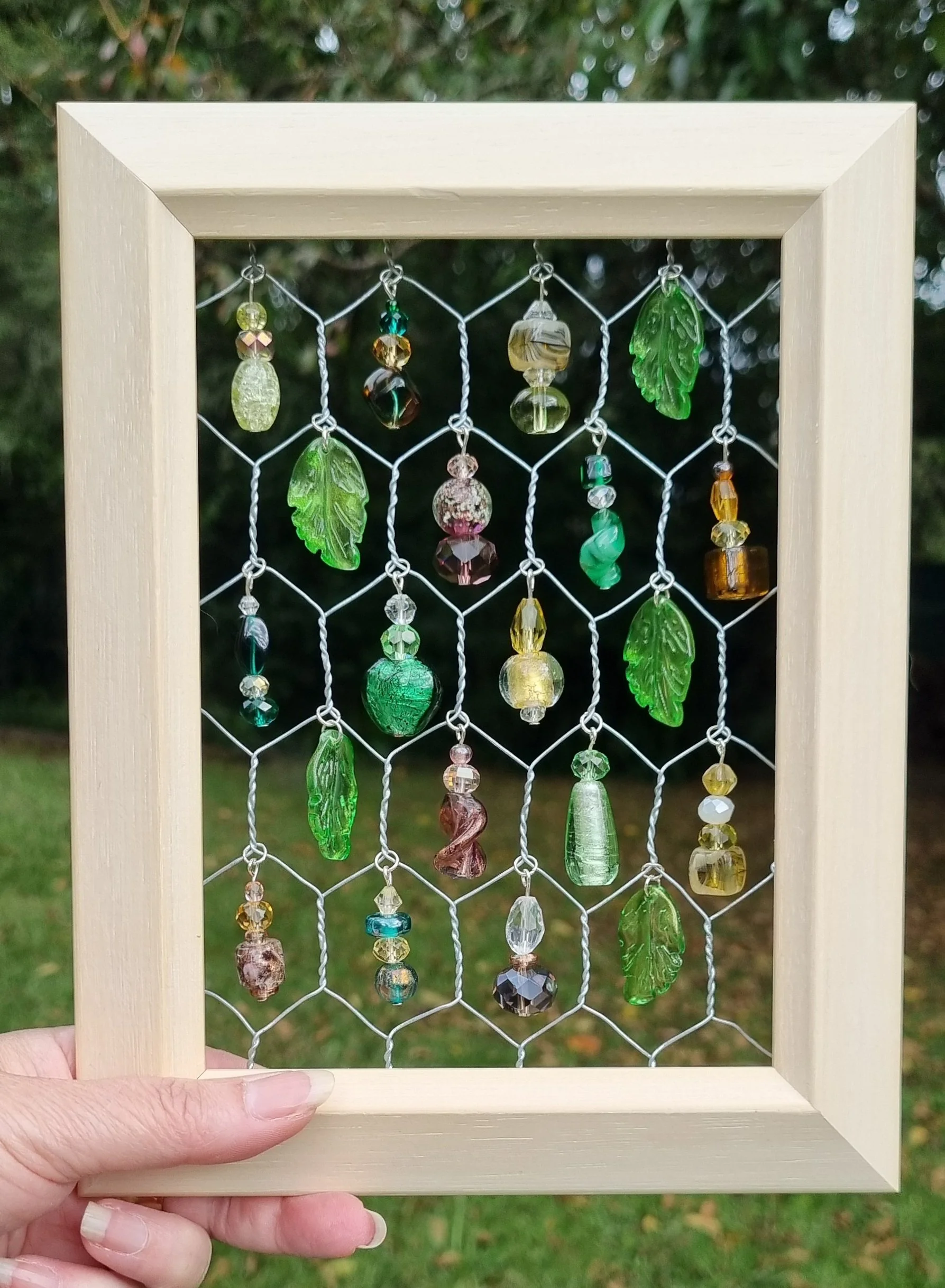 Hand holding a wooden frame with wire mesh decorated with green glass leaf-shaped beads, various other colorful glass beads, and stones, outdoors with trees in the background.