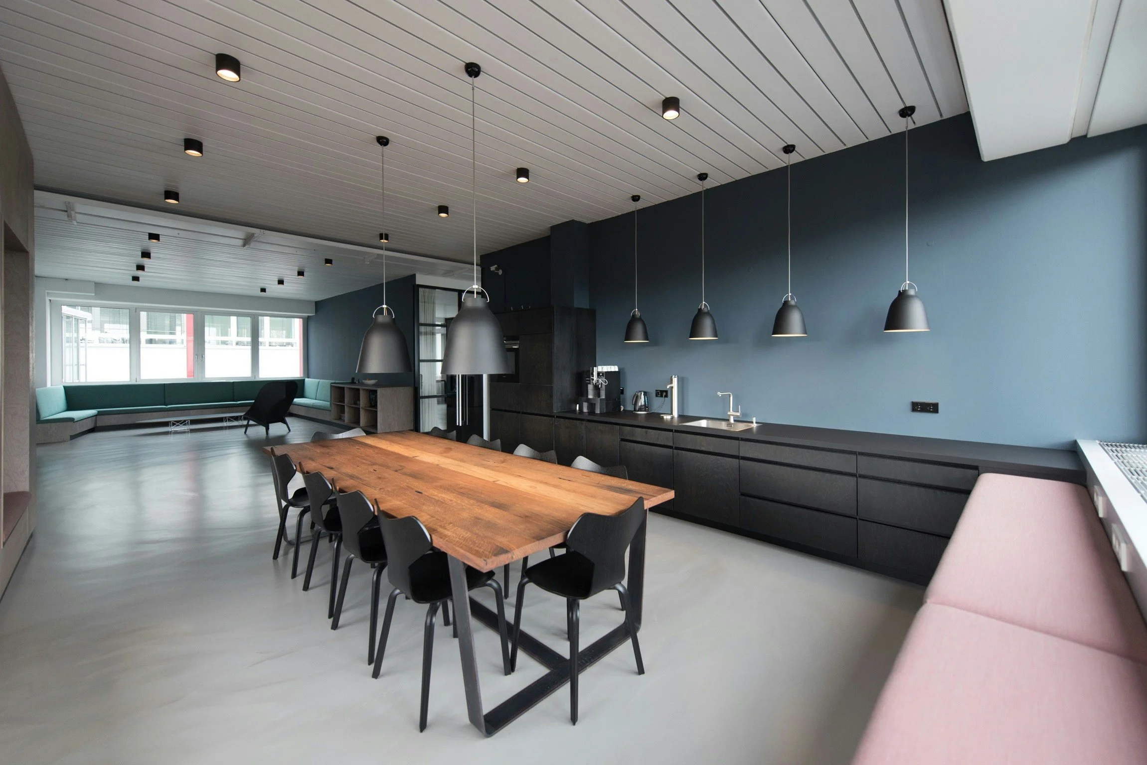 Modern kitchen and dining area with black pendant lights above a large wooden table, black chairs, and a blue accent wall with built-in black cabinets and a coffee station, adjacent to a lounge with teal and black seating and large windows.