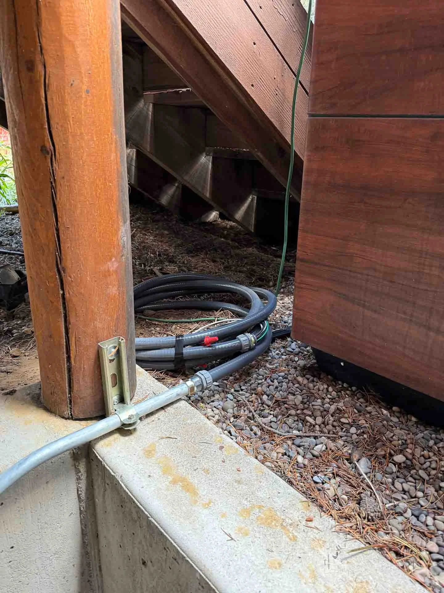 Close-up of a corner of an outdoor wooden structure showing electrical conduit, hoses, and gravel ground.