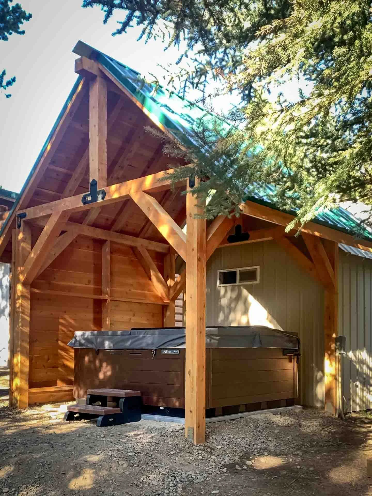 A hot tub with a brown exterior situated under a wooden pergola with a green metal roof, with trees and sunlight filtering through branches.
