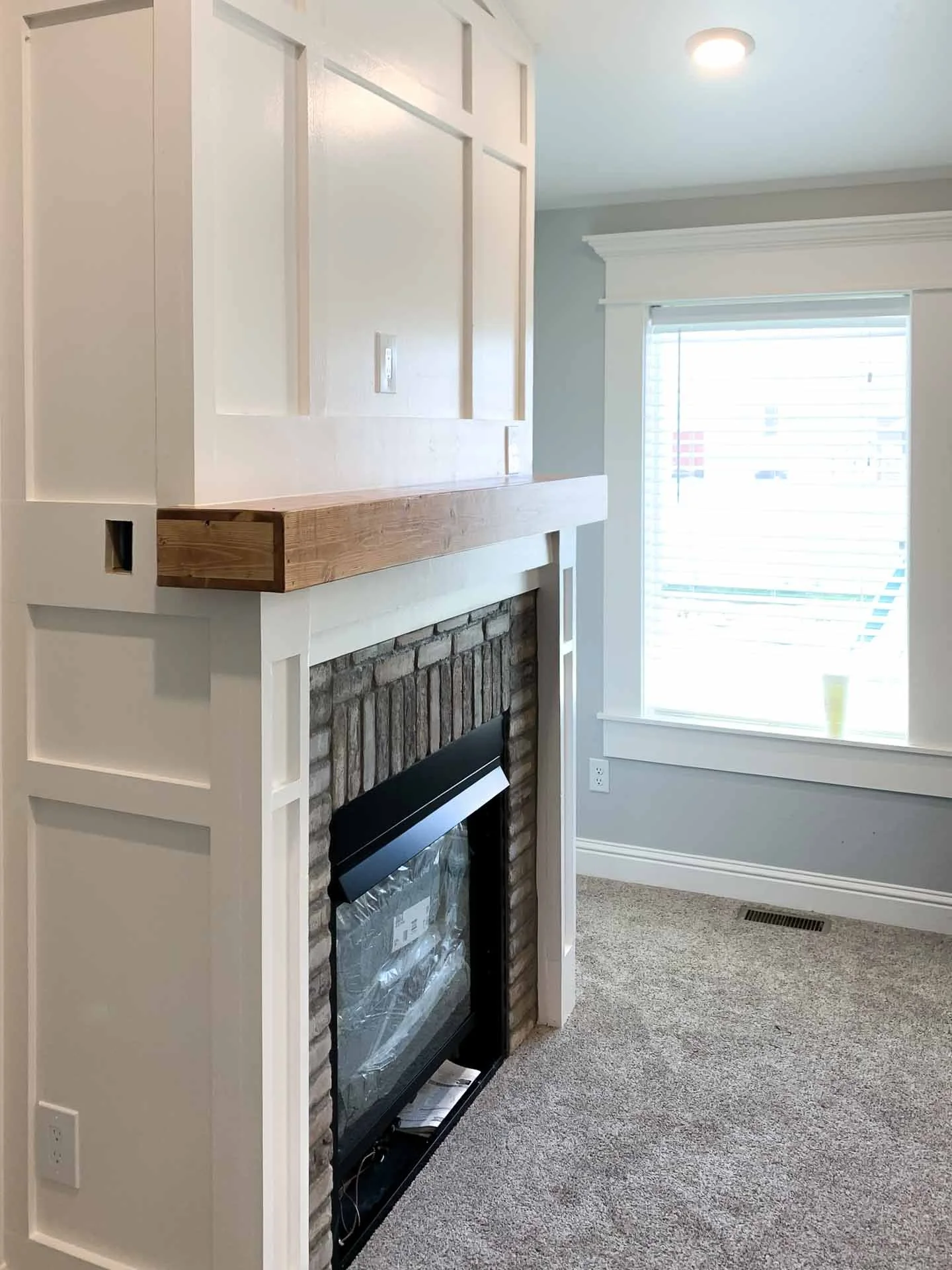 A room with a partially built white-painted fireplace with wood trim and brick surround, located next to a window with white blinds and trim, and beige carpeted floor.