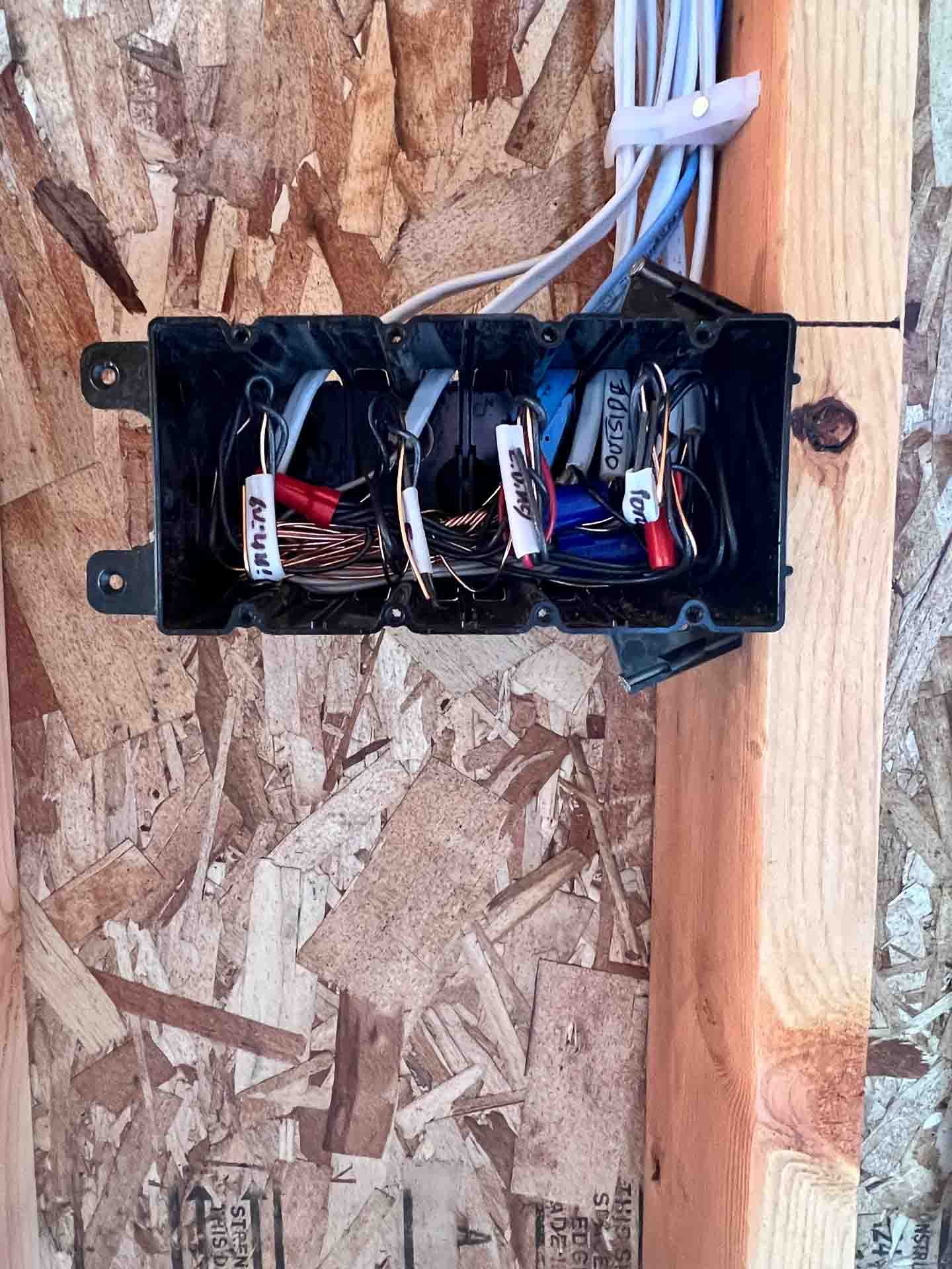 An open black plastic electrical junction box with wires inside, labeled with white tags, mounted on a wooden surface with a wood piece nearby.