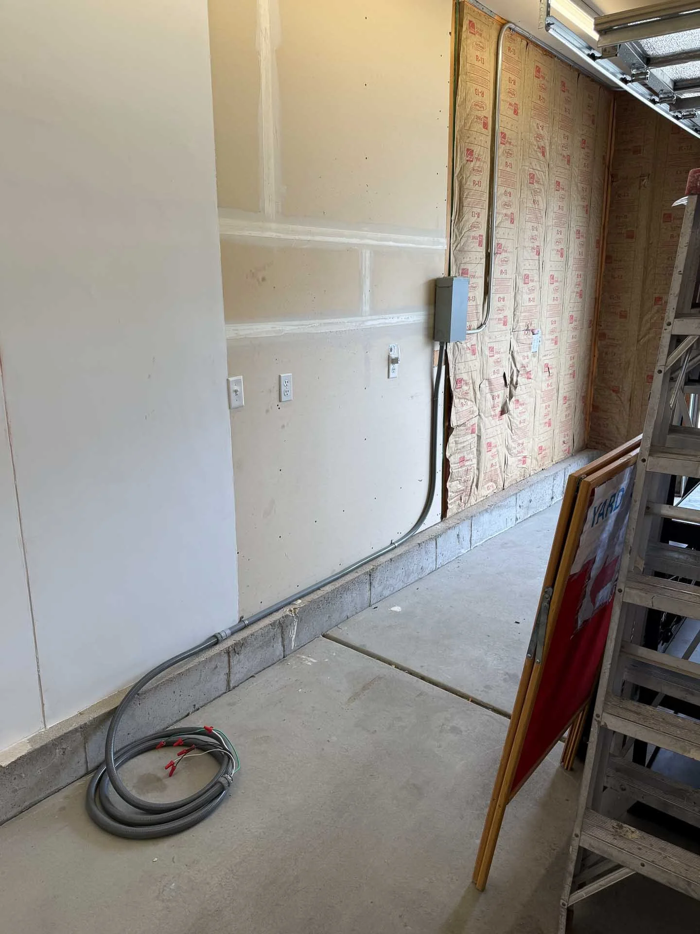 Construction site showing an unfinished garage wall with electrical outlets, conduit piping on the floor, exposed insulation on one wall, and a ladder and construction sign on the right side.