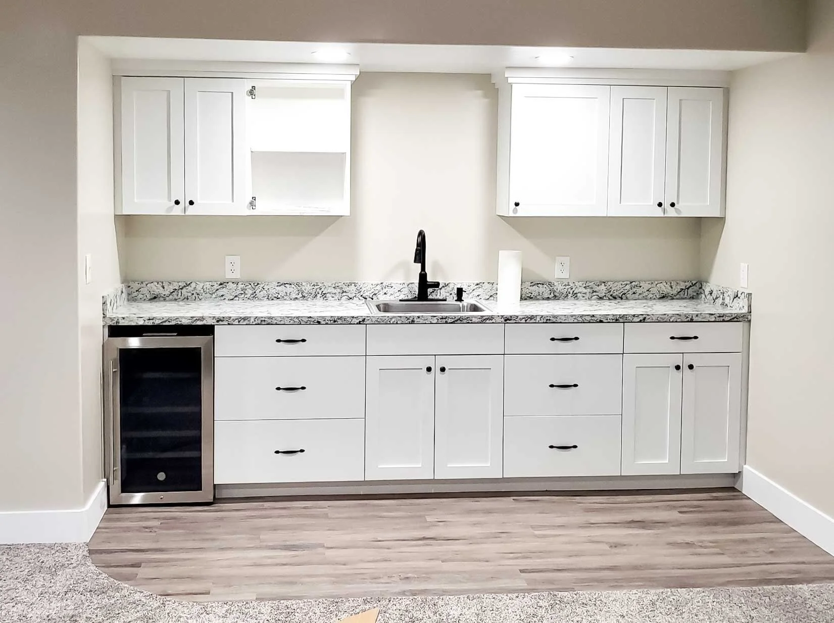 Kitchen with white cabinets, granite countertop, black faucet, small wine cooler, and wooden flooring.