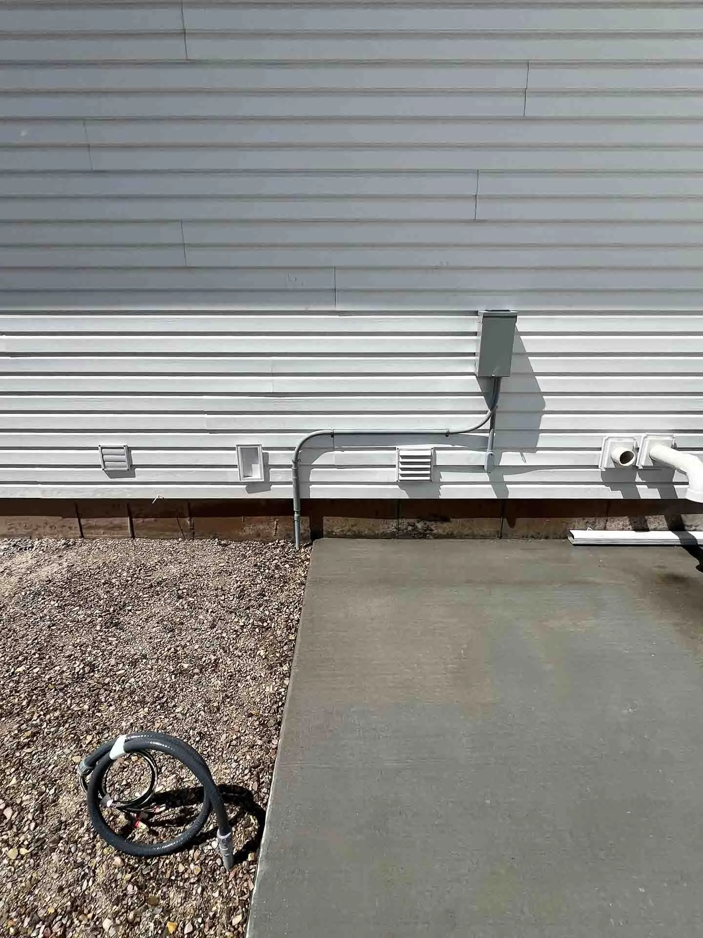 Exterior wall of a building with white siding, electrical conduit, vents, and drainage pipes, along with a coiled outdoor electrical cord and concrete walkway.