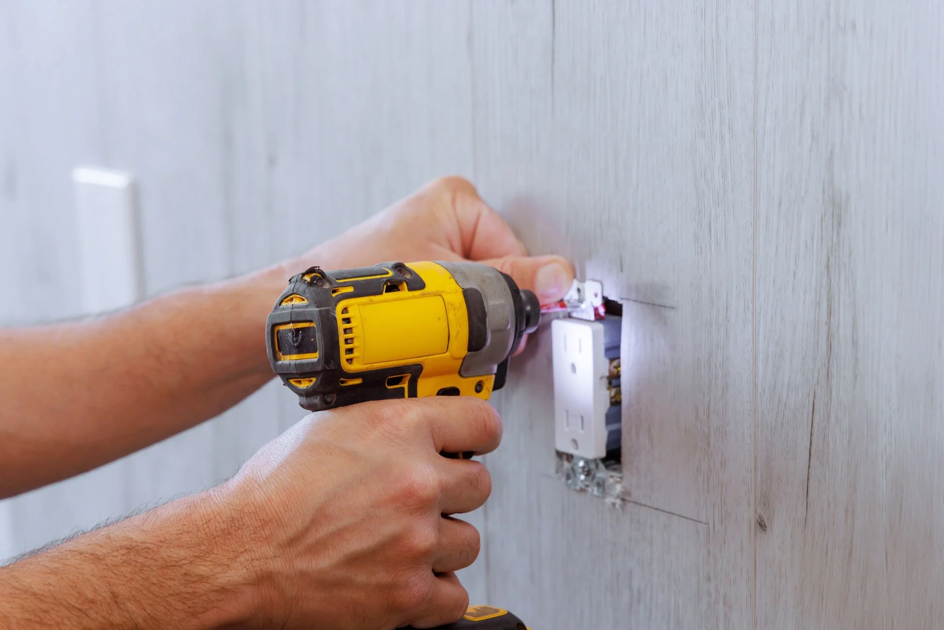 A person using a yellow cordless drill to work on an electrical outlet in a wall, with the outlet cover removed.