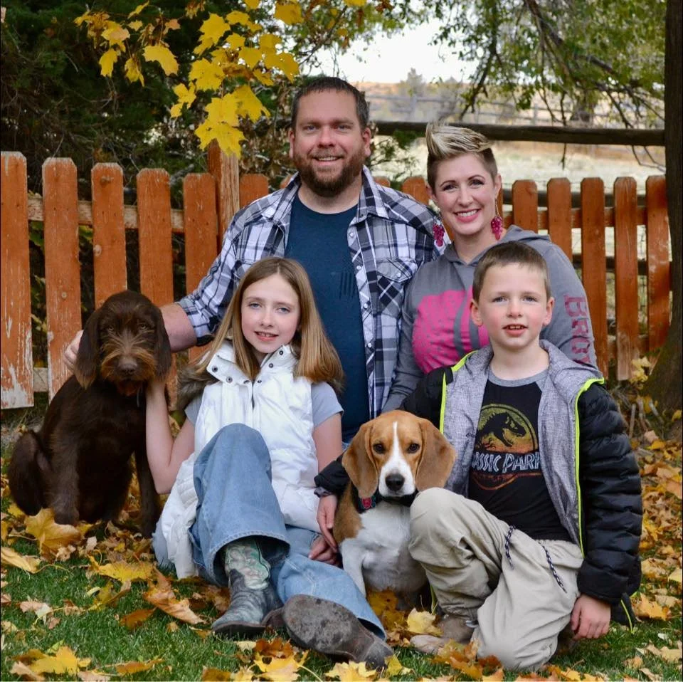 A family of four with two dogs stands outdoors in autumn surrounded by fallen leaves, a wooden fence, and trees with yellow leaves. The father has short dark hair and a beard, wearing a plaid shirt. The mother has short blonde hair and is wearing headphones around her neck. The daughter has long blonde hair, wearing a white vest and jeans. The son has short light brown hair, wearing a Jurassic Park shirt and a gray jacket. The two dogs are a brown puppy and a beagle, which the children are holding or sitting next to.