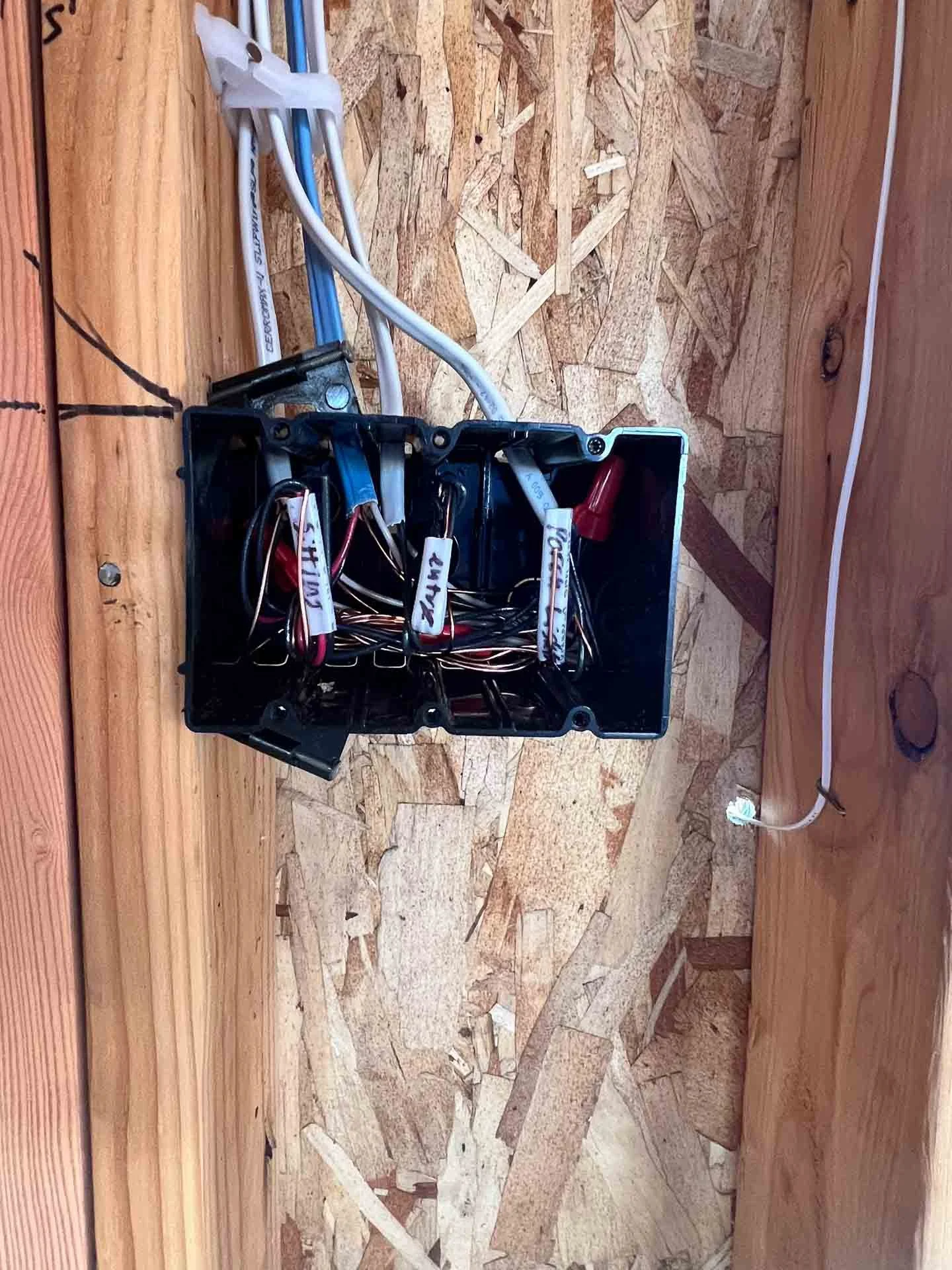 Open black plastic electrical junction box with wires inside and labels, mounted on a wooden surface with a white power cord plugged into a wall outlet.
