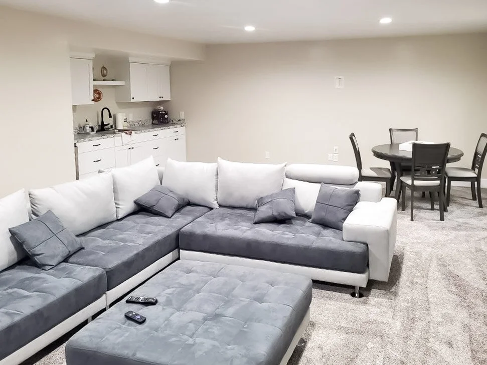 Living room with white and gray sectional sofa, a TV remote on the ottoman, kitchen area in the background, and a dining table with four chairs to the right.
