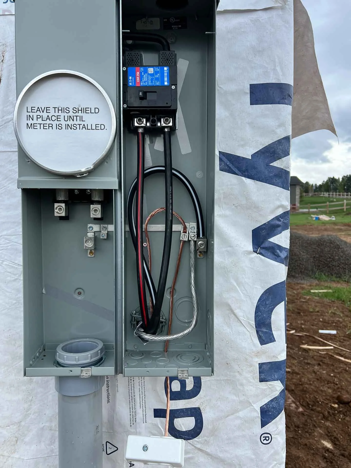 An electrical meter box with wiring inside, a label that says 'Leave this shield in place until meter is installed', and a partially visible utility pole outside.