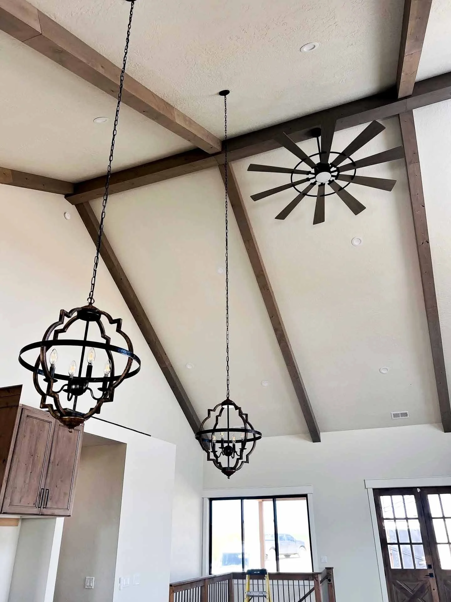 Interior view of a room with a high ceiling featuring exposed wooden beams, two ornate hanging light fixtures, a ceiling fan, and large windows with a staircase railing in the background.
