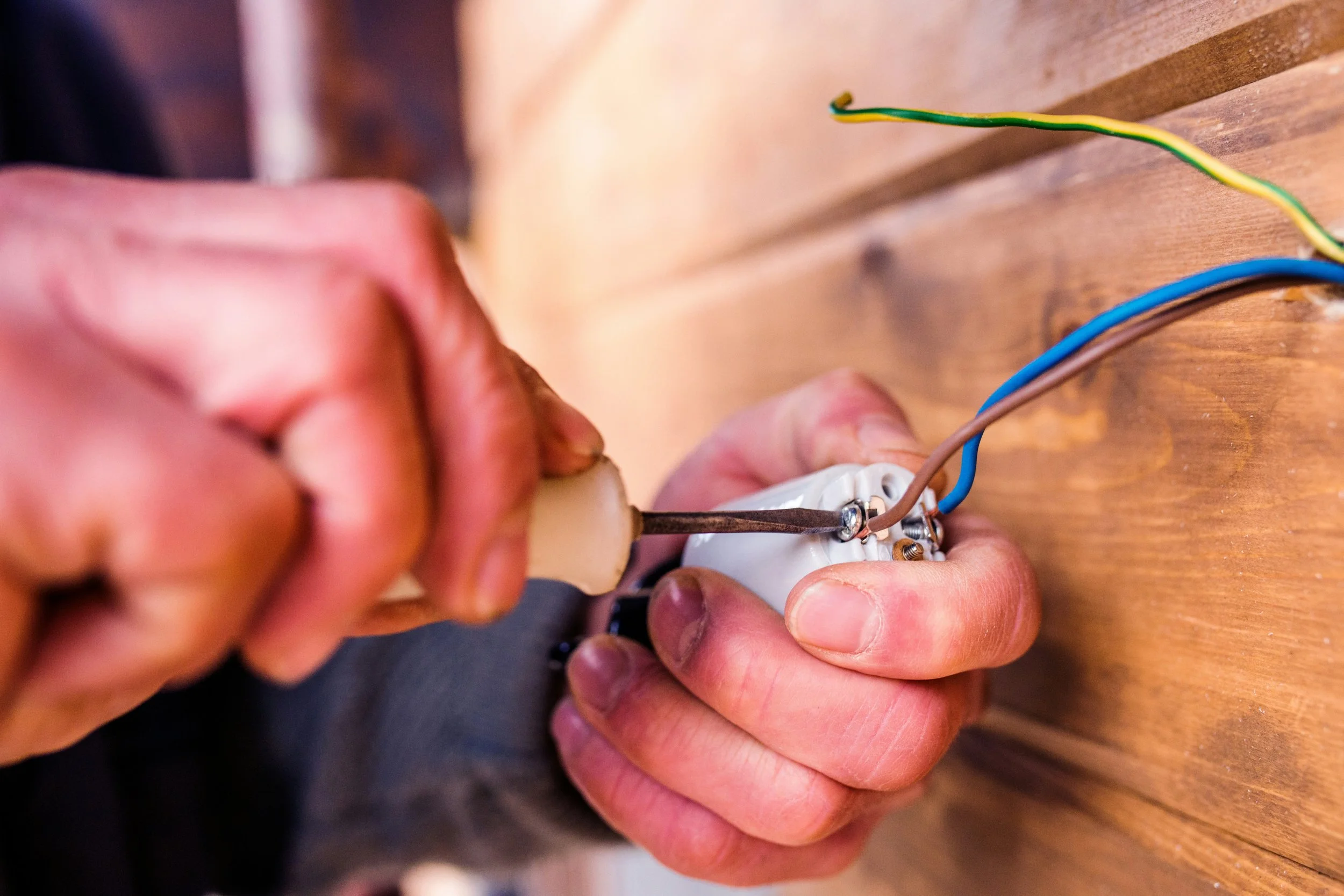 A person working on wiring an electrical outlet, using a screwdriver to connect wires to an electrical socket, with several wires visible on a wooden surface.