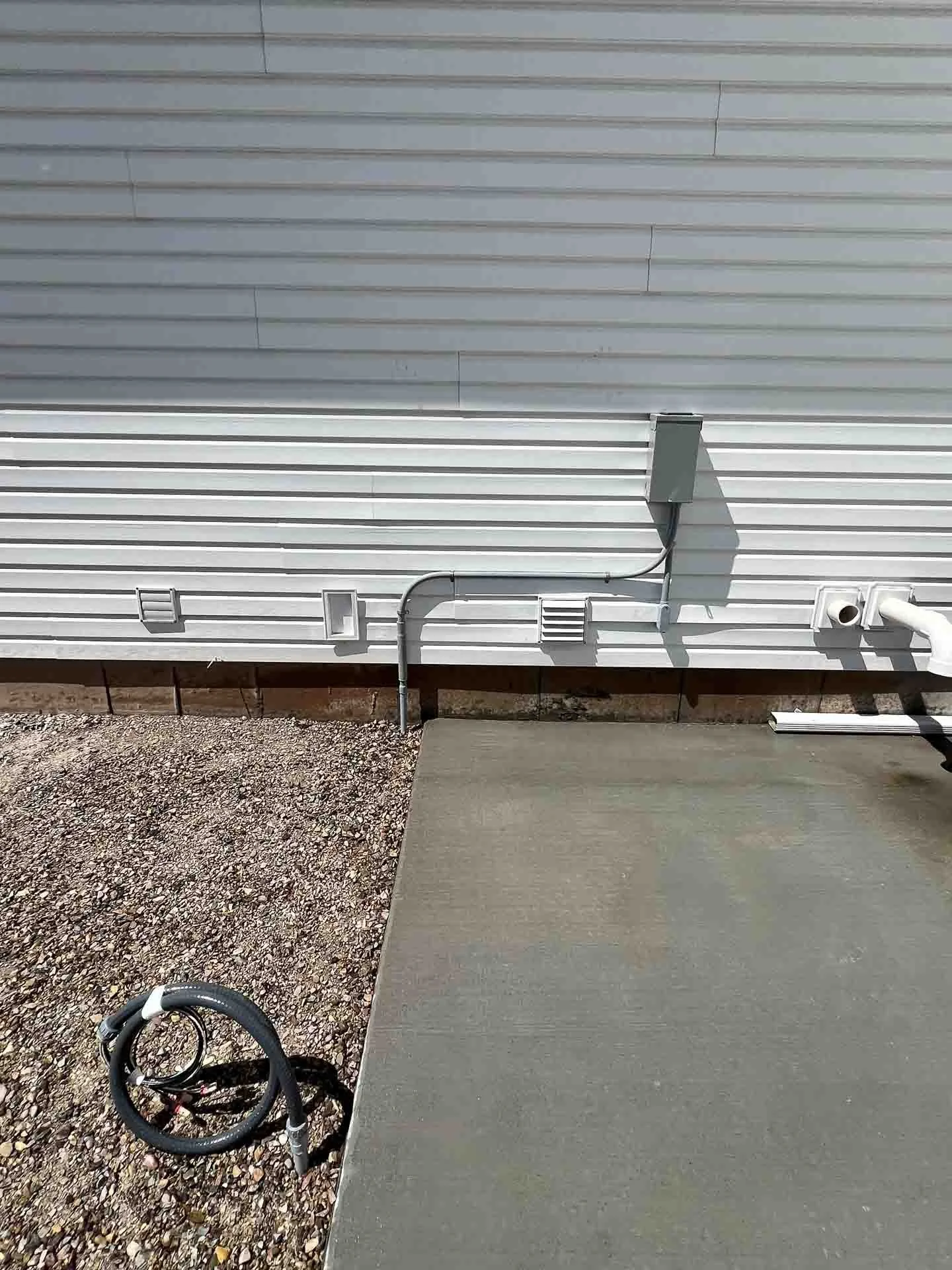 Outside view of a house wall with electrical wires, cables, and ventilation openings, next to a concrete slab and gravel ground.