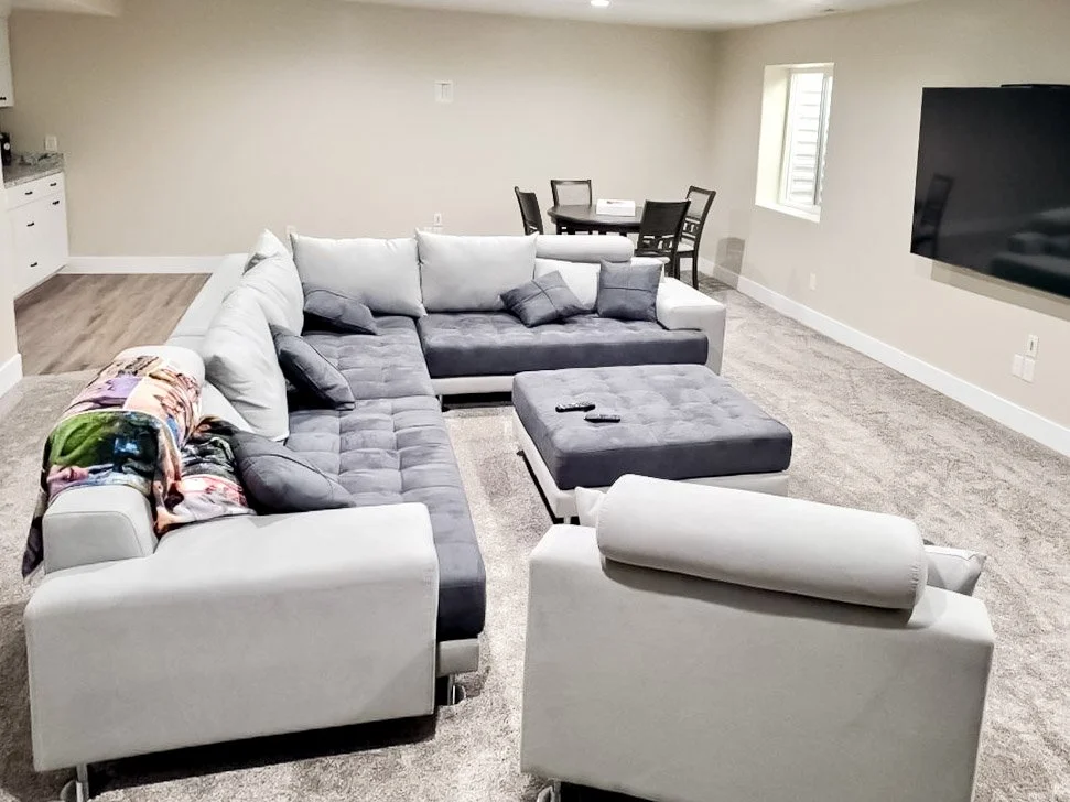 Living room with a sectional sofa, an ottoman, armchair, a wall-mounted TV, a dining table with chairs, and a window.