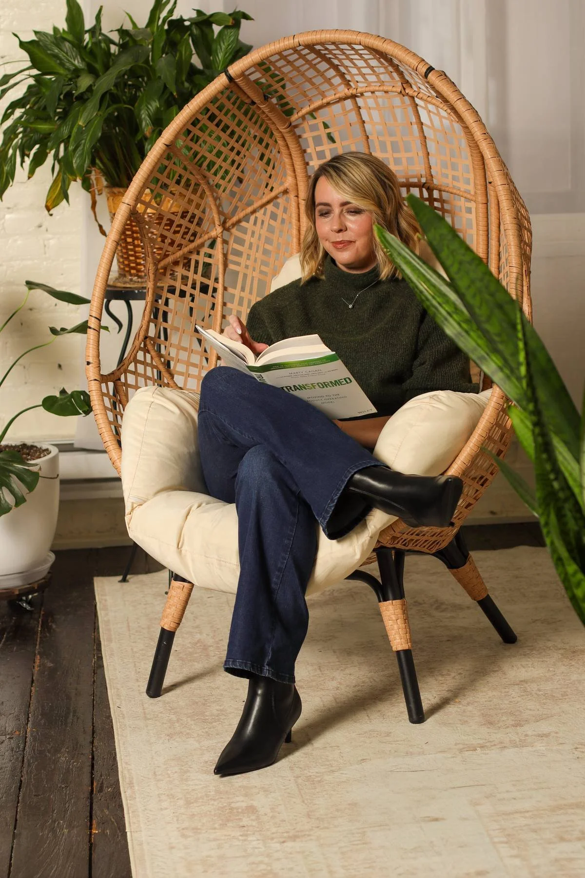 A woman with blonde hair wearing a green sweater, jeans, and black boots sitting in a rattan hanging chair, reading a book titled "Transformed," in a cozy indoor setting with green plants.