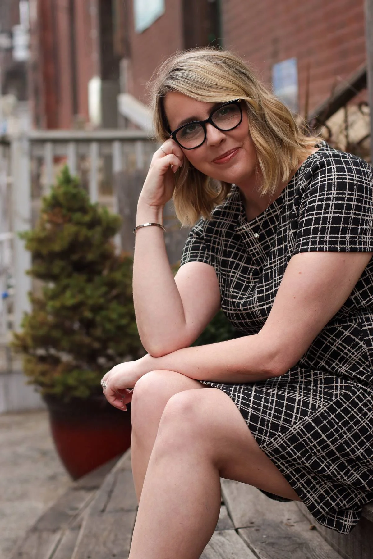 A woman with blonde hair and glasses sitting outdoors on a wooden bench, wearing a black and white checkered dress, with potted plants and brick buildings in the background.