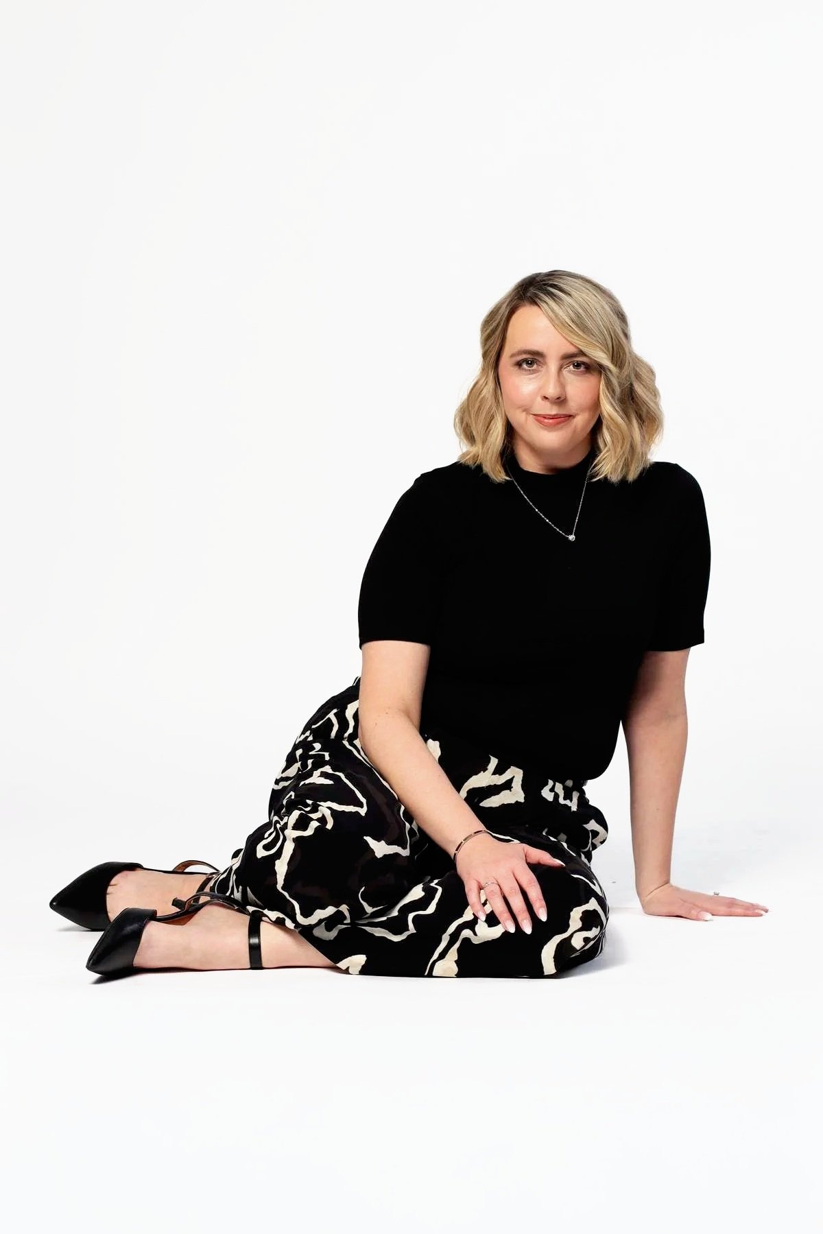 A woman with shoulder-length blonde hair is sitting on the white floor, wearing a black short-sleeve top, a black and white patterned skirt, and black high-heeled shoes. She is smiling softly and looking at the camera.