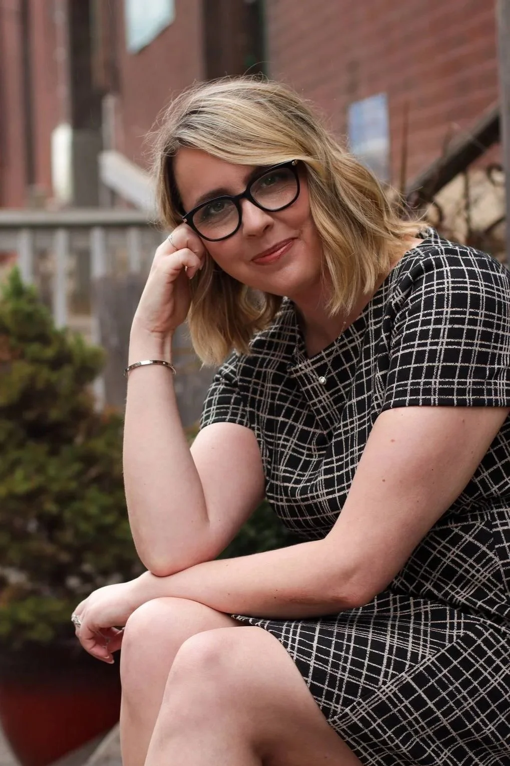 A woman with blonde hair, glasses, and light skin sits outdoors, resting her head on her hand, wearing a black and white checkered dress.