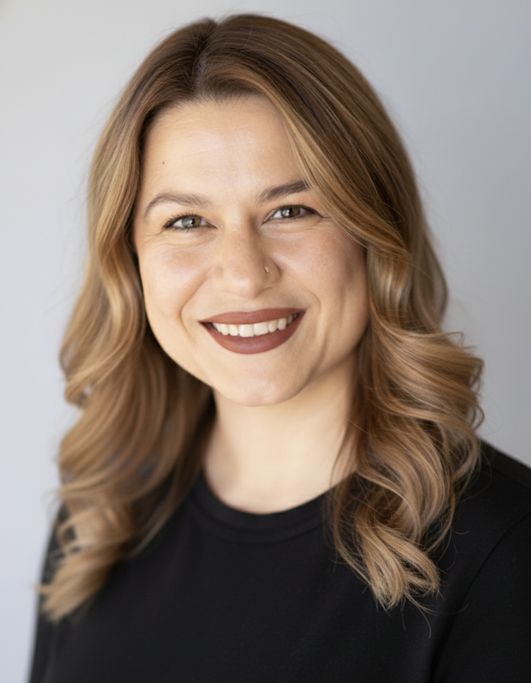 Portrait of a woman with wavy, reddish-brown hair, wearing a black top, smiling with visible teeth, and light makeup.