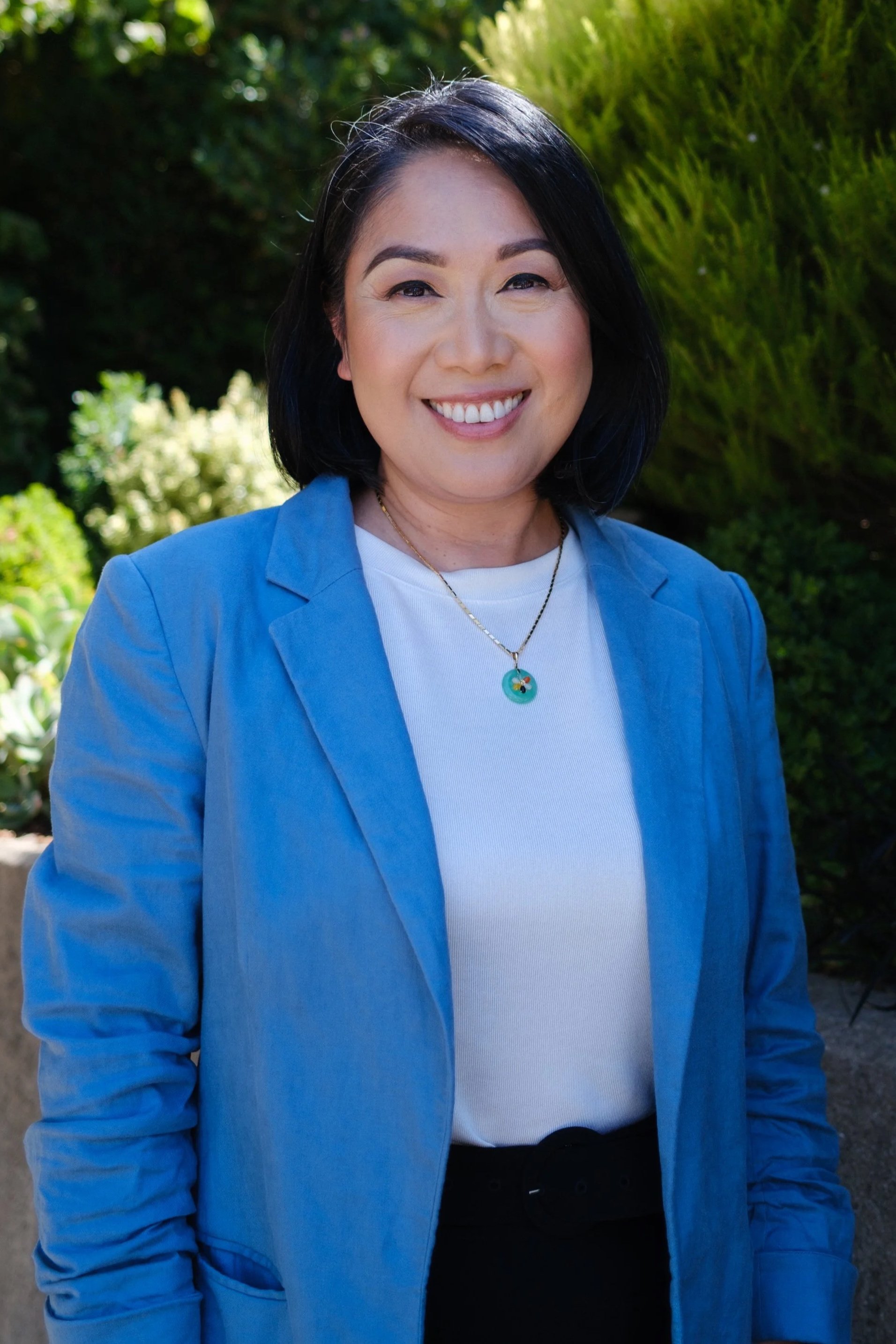 A woman with shoulder-length black hair, wearing a light blue blazer over a white top, standing outdoors with greenery in the background.