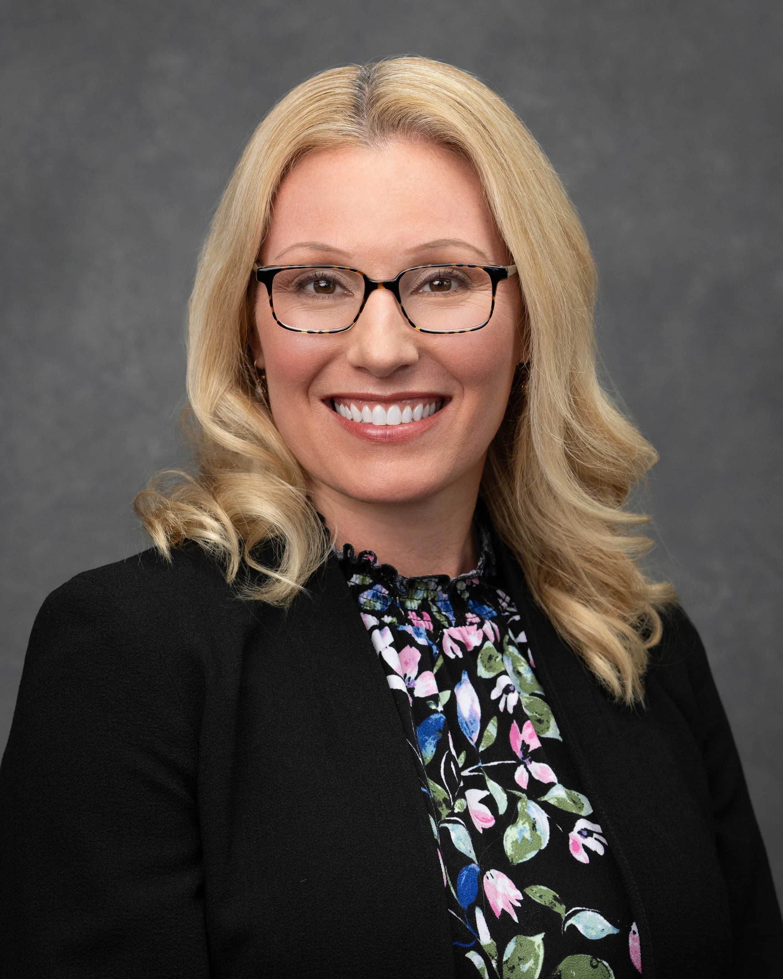 Close-up of a woman with blonde hair and glasses, smiling against a gray background