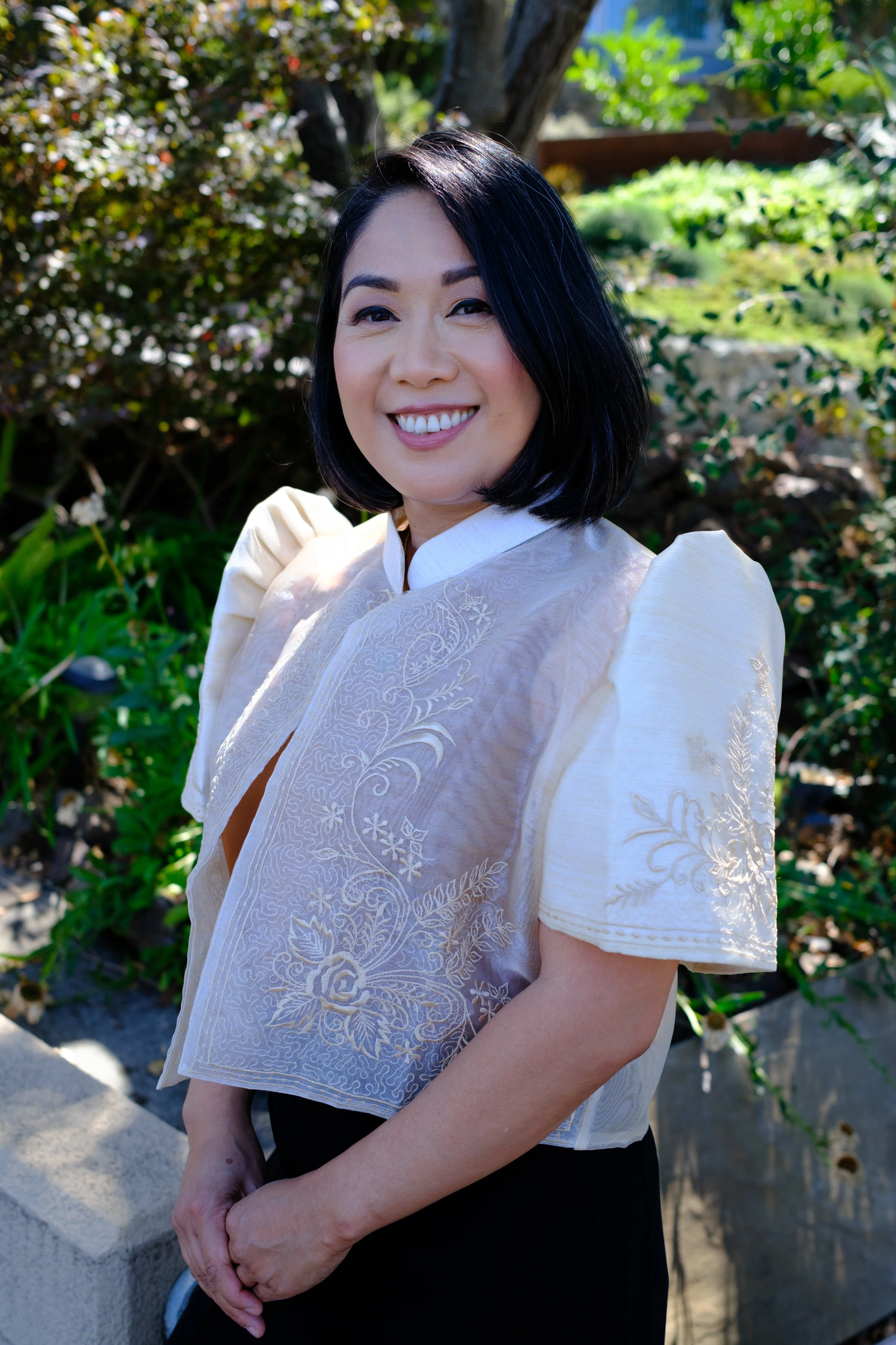 A woman with short black hair smiling outdoors, wearing a light-colored, embroidered blouse and black pants, with greenery and a tree in the background.