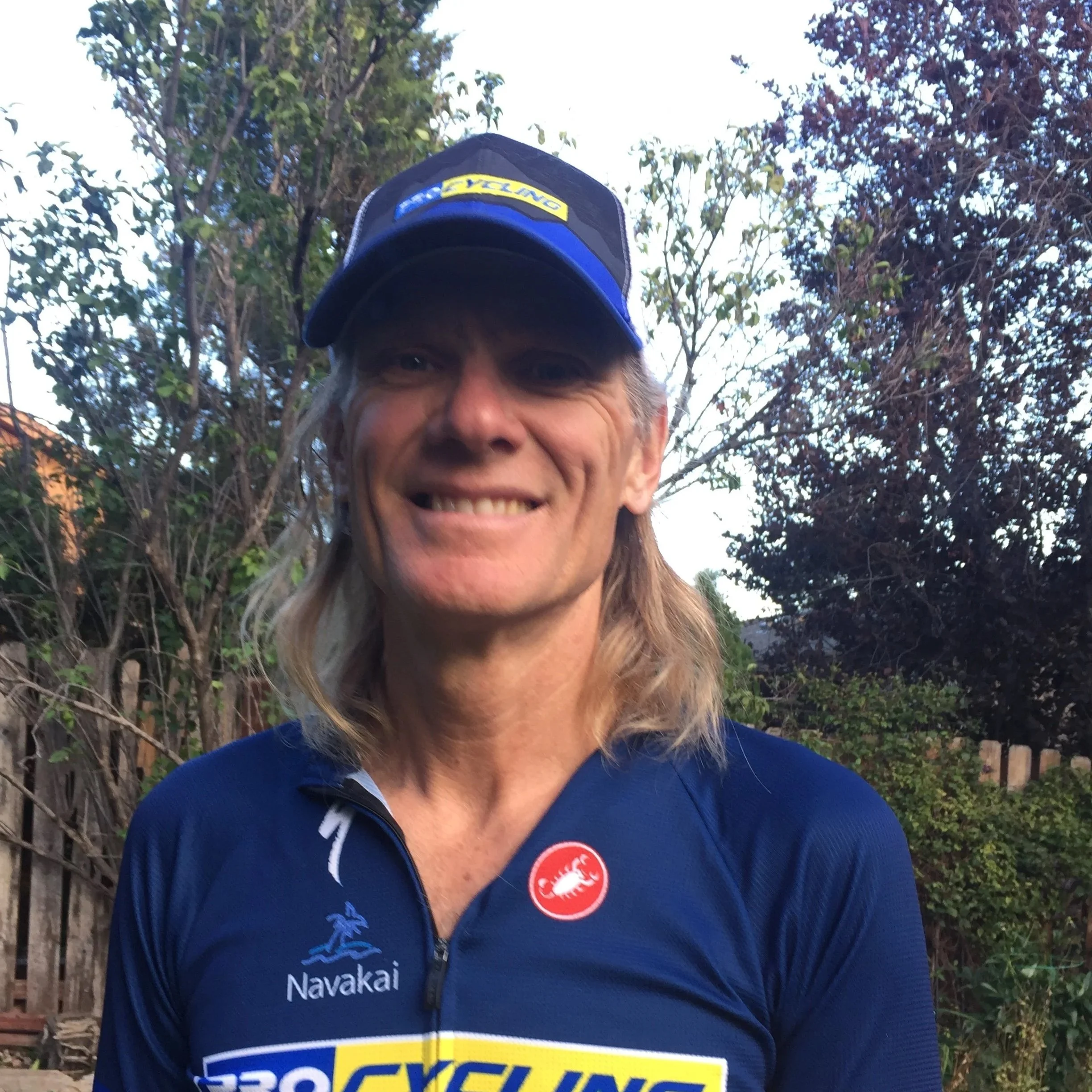 A smiling man with long blonde hair wearing a blue cycling jersey and a blue cap, standing outdoors with trees and a fence in the background.