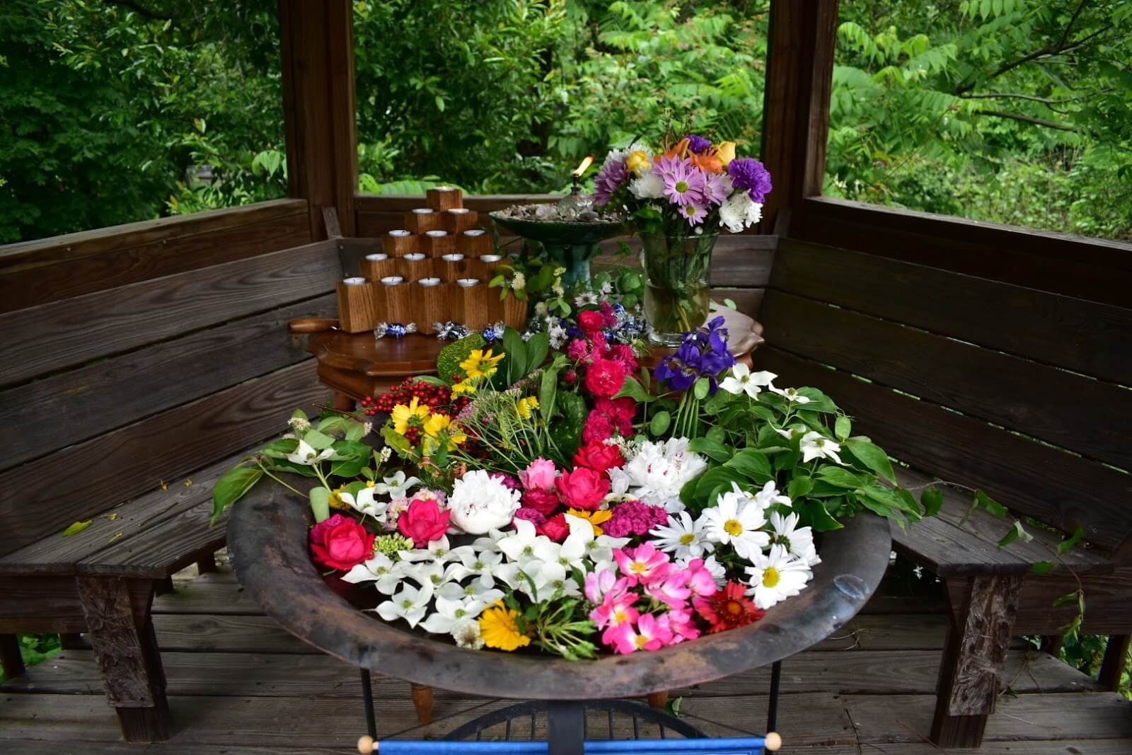 A round table topped with assorted colorful flowers sits in a wooden gazebo, with more flowers and wooden candle holders arranged in the background. Dense greenery is visible outside.