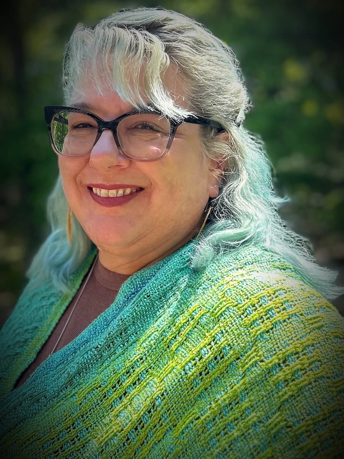 A portrait of Reverend Mandy Goheen, a smiling woman with grey and teal hair, wearing black-framed glasses and a textured green and yellow woven shawl against a soft-focus garden background.