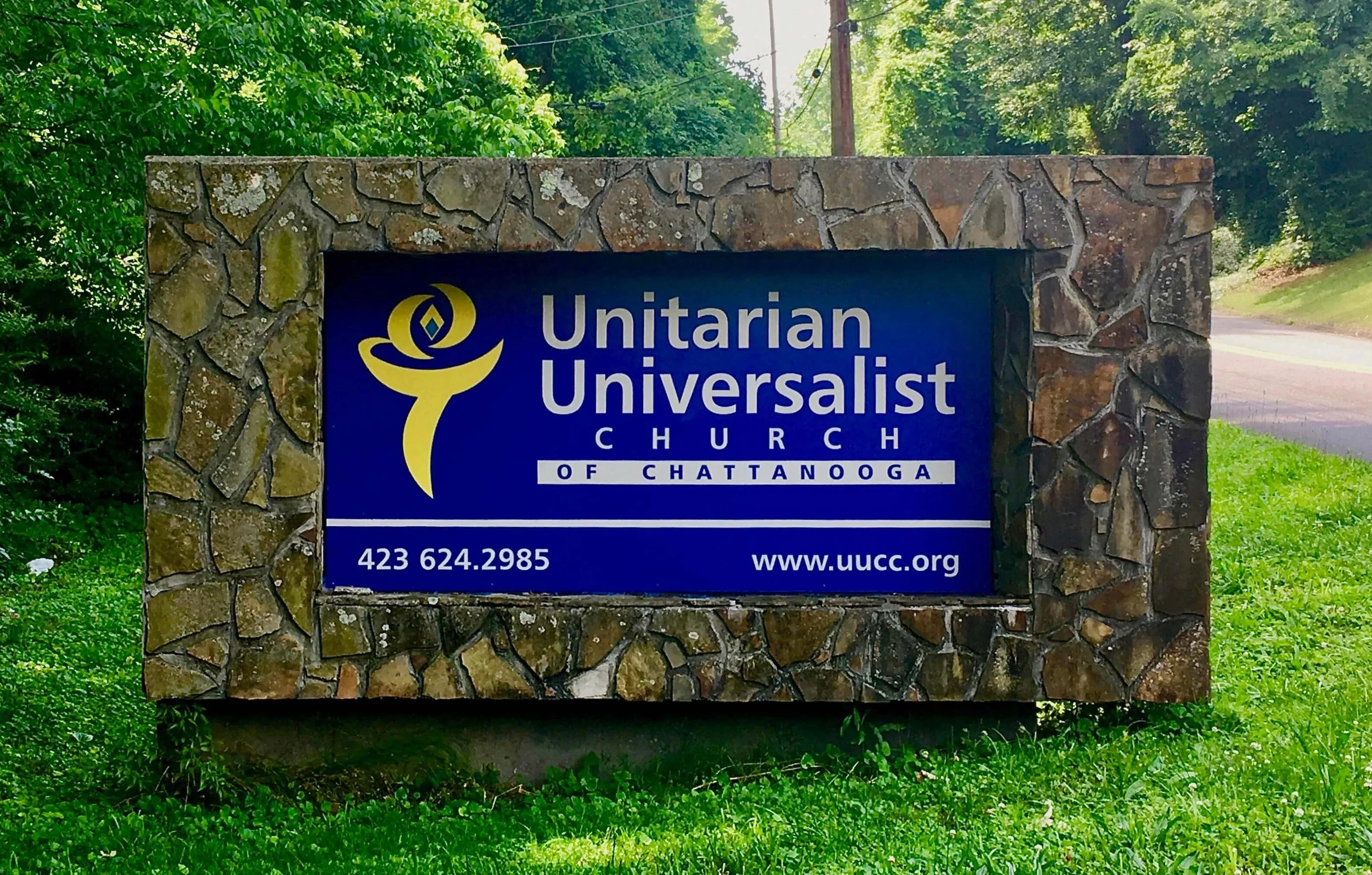 A roadside sign for the Unitarian Unversalist Church of Chattanooga. The sign is set in a rustic stone frame and features a blue background with a yellow flaming chalice logo. Text includes the church, name, phone # 423-624-2985, and www.uucc.org.