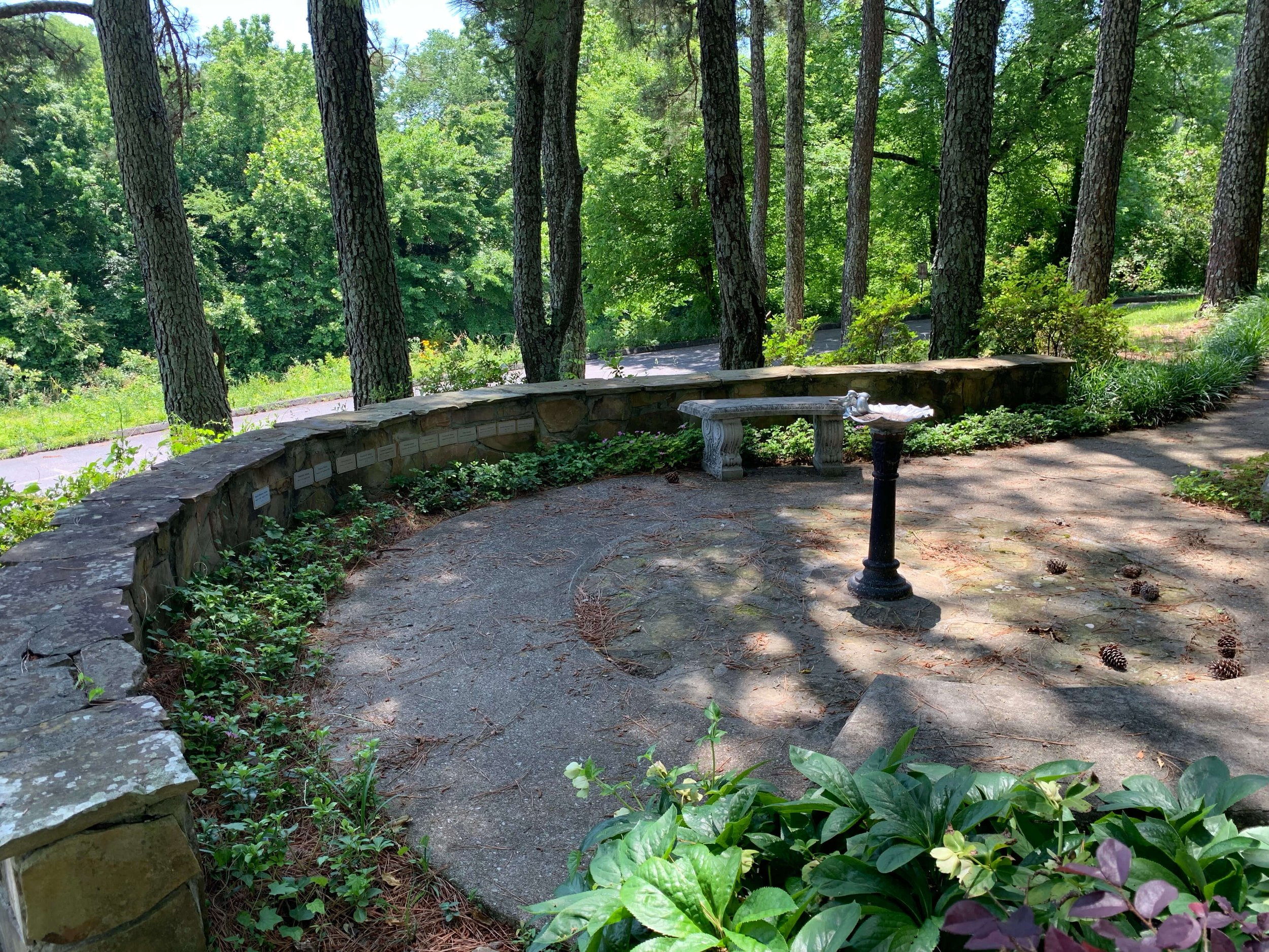A peaceful church memorial garden featuring a curved stone wall with small memorial plaques, a grey stone bench, and a dark pedestal birdbath, all set under the shade tall trees and lush greenery.