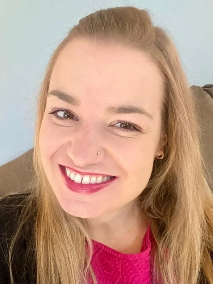 Close-up of a woman with long gingery blonde hair, smiling at the camera, wearing a bright pink top and a dark jacket, sitting on a brown couch with a light blue wall in the background.