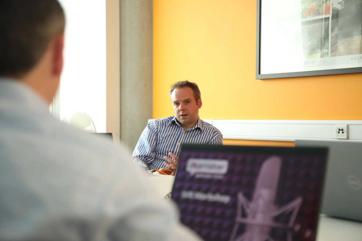 Two men in a meeting room, one facing the camera with a serious expression, while the other has his back to the camera. The man facing the camera is wearing a striped shirt. There are laptops on the table, and a yellow wall with a framed picture.