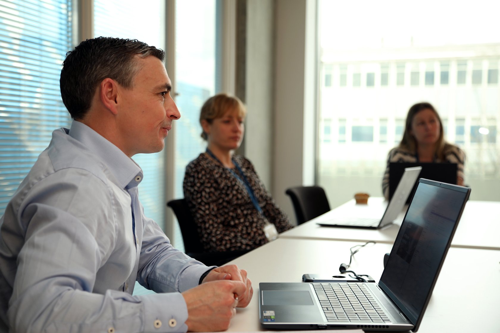A man and two women sitting at a conference table in an office, with laptops in front of them, during a meeting.