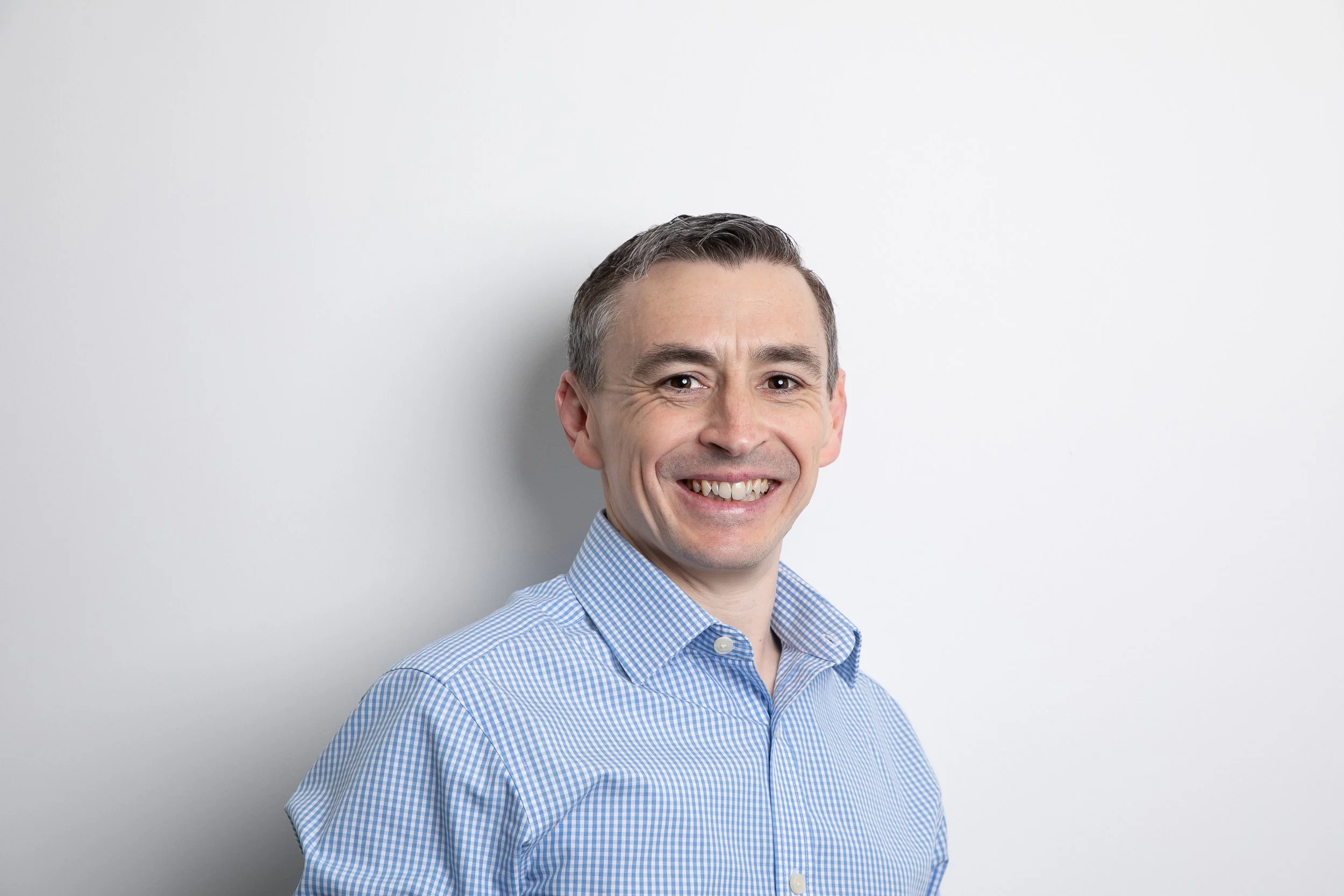 A middle-aged man with short dark hair, smiling, wearing a light blue checkered shirt, standing against a plain white background.