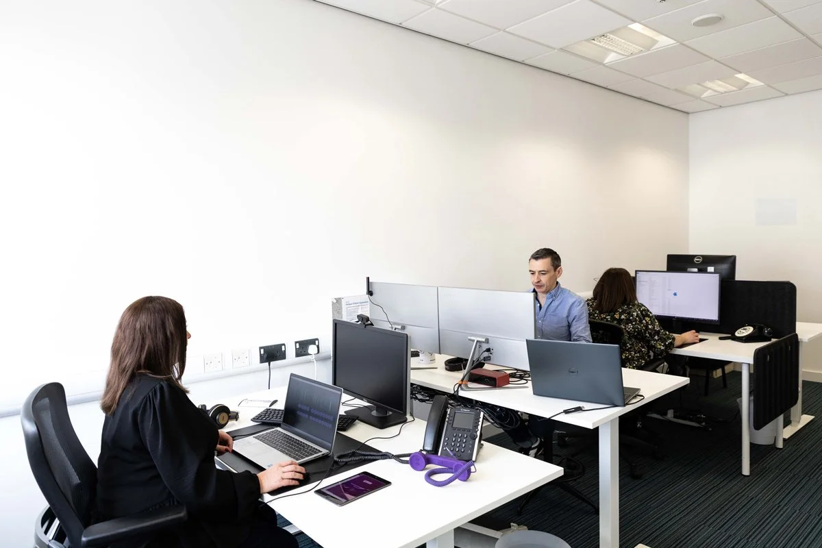 Office workers working at desks in an open-plan office with multiple computer monitors, phones, and office supplies.