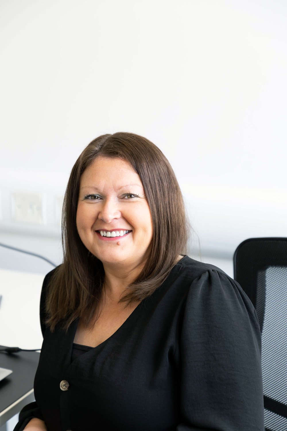 A woman with brown hair smiling at the camera, sitting in an office chair, wearing a black top, with a white wall in the background.